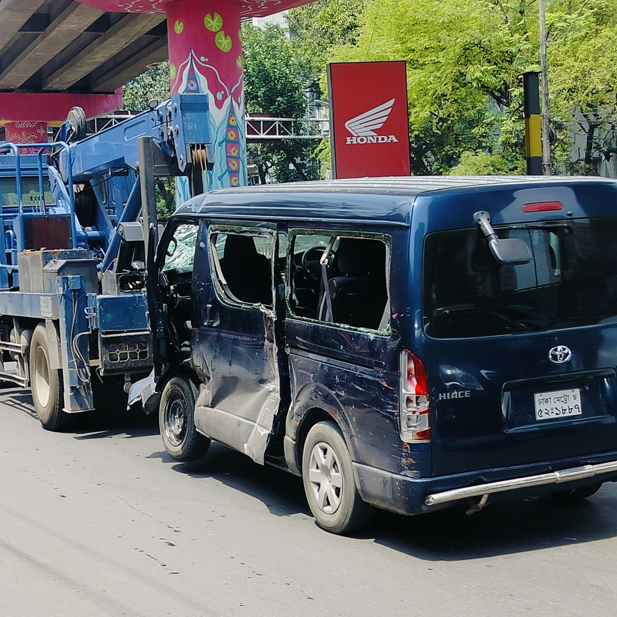 A damaged blue Toyota HiAce van is being towed by a blue crane truck in Dhaka, Bangladesh. The van's windows are broken, and there is extensive damage to the driver's side, including the door. The van's license plate reads "ঢাকা মেট্রো-চ ৫২-১৮৮৭". In the background, there is a Honda sign and a bridge support painted with floral designs. Trees are visible in the background, indicating an urban environment. The scene suggests a recent accident.