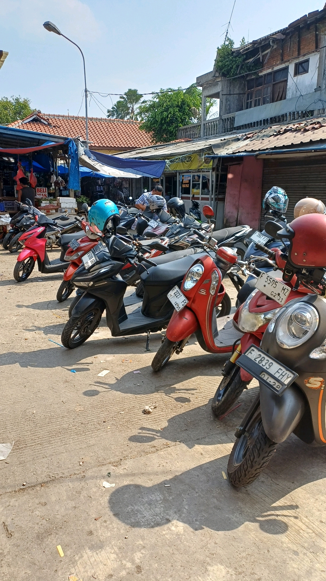 In an outdoor scene in Citeureup, Indonesia, multiple parked scooters are lined up on a concrete surface under a clear sky. A lamppost stands tall on the left, casting a shadow. Several stalls with red-tiled roofs and blue tarps are visible in the background, suggesting a market or commercial area. One stall displays goods and a sign reading "BELI MAS" (Buy Gold). Another stall has windows and signage, possibly indicating food or services.
A variety of scooters are prominently featured, many with helmets placed on them. The scooters are of different colors, including red, black, and pink. License plates are visible on some of the scooters, with readable plates showing characters such as "F 2227 FS", "F 5505 MZ", and "F 2839 FHY". One scooter has a blue helmet on its handlebars, while others have helmets with various designs. In the background, two men are partially visible, one standing and looking towards the stalls and another seated on a scooter. The ground is paved with concrete and scattered with some debris, including paper scraps and plastic. The overall impression is a typical street scene in a bustling Indonesian town.