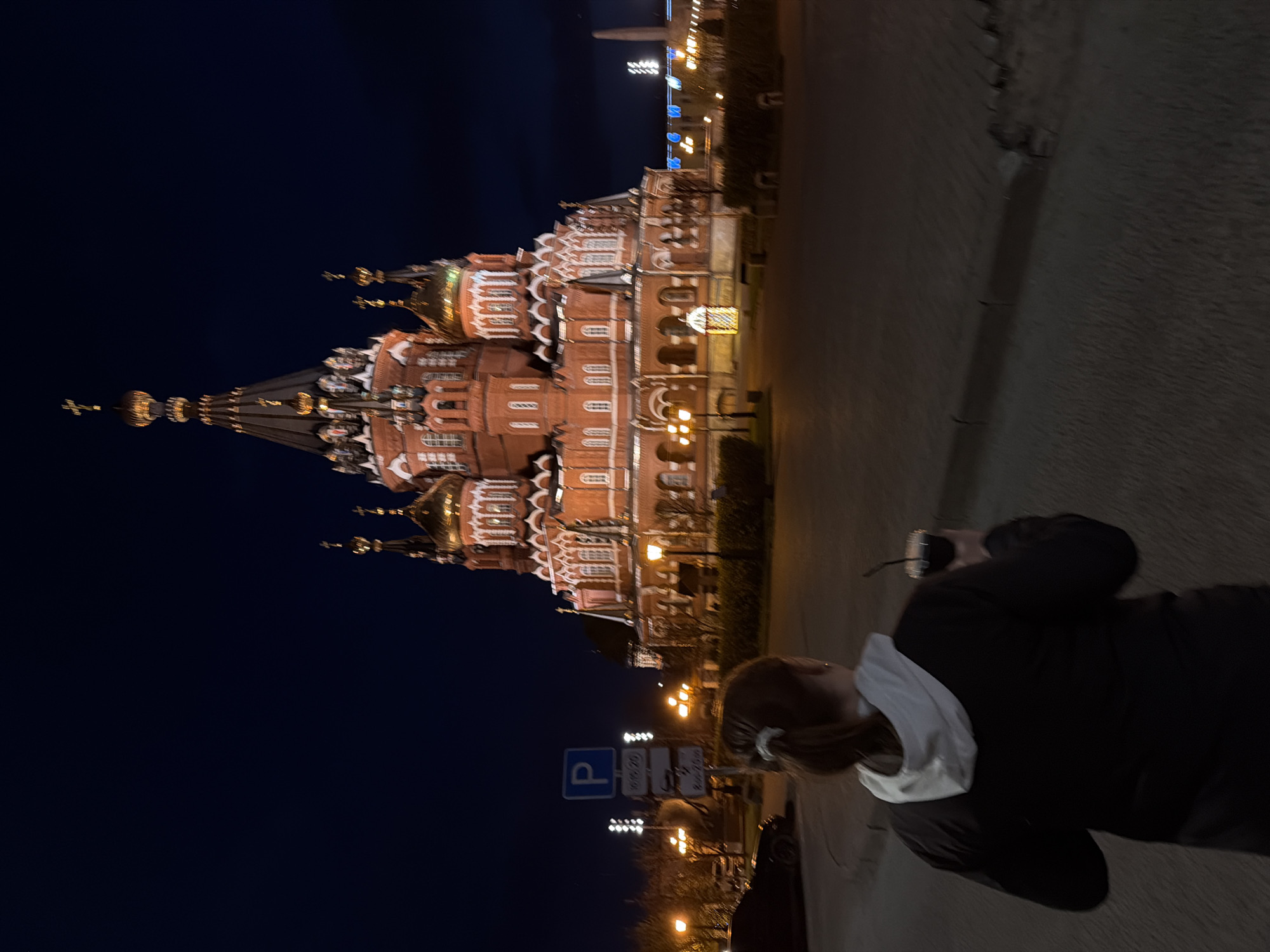 A nighttime shot shows the brightly lit St. Michael's Cathedral in Izhevsk, Russia. To the right of the church is a paved sidewalk with a person walking on it. The person is wearing a black jacket with a white collar and has light brown hair pulled back. Parking signs are visible near the church.