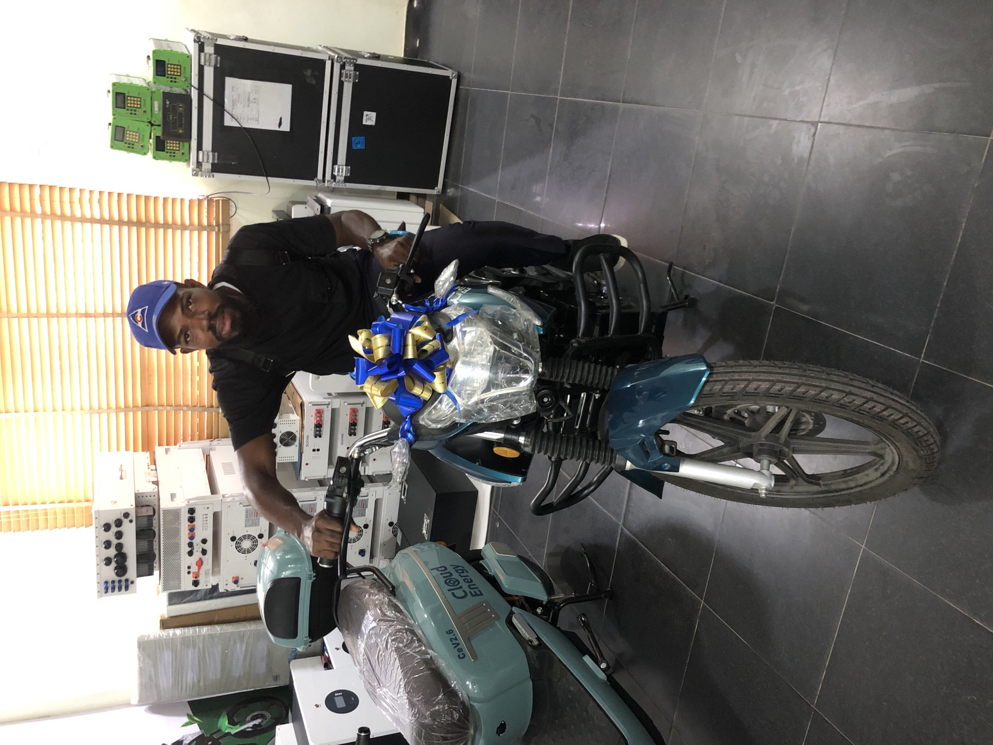 A man wearing a blue baseball cap and a black t-shirt is sitting on a teal electric motorcycle in a room that appears to be a showroom or workshop. The motorcycle is decorated with a blue and gold bow on the handlebars. Behind the man, there are stacks of electronic equipment, including inverters and power systems, and a large black flight case. To the left of the motorcycle, another teal electric scooter is partially visible. The room has tiled flooring and is illuminated by natural light filtering through blinds on a window in the background. The overall impression is a professional setting likely related to energy solutions, possibly located in Ikeja, Nigeria, given the "Cloud Energy" branding on the scooter and the prevalence of such businesses in the area.