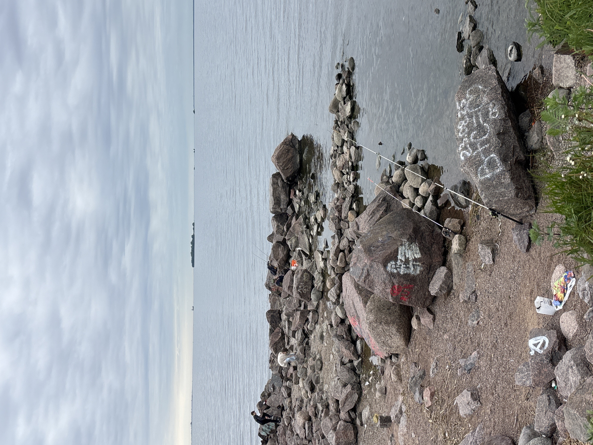 The scene depicts a rocky shoreline along the Gulf of Finland, situated in the city of Saint Petersburg, Russia. The sky is overcast and the water is a pale grey. Several people are on the rocks, some fishing. Fishing rods are propped up against the rocks. The ground near the edge of the rocks is littered with trash. Graffiti is visible on some of the rocks. In the distance, there's a thin strip of land on the horizon.