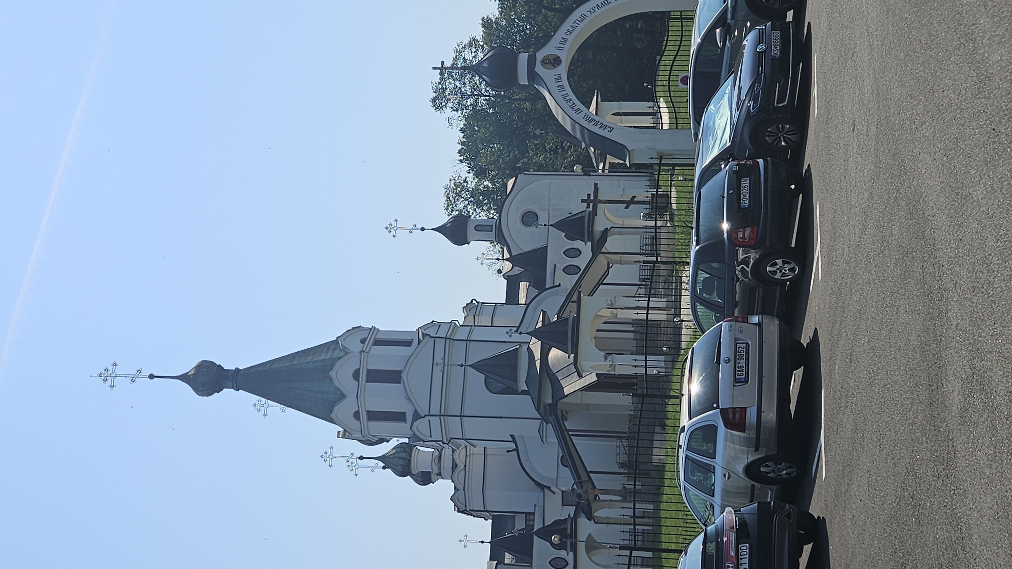 The image shows the Holy Trinity Orthodox Church in Prešov, Slovakia. The church is a white building with multiple domes topped with crosses. In front of the church, there is a parking lot with several cars. The sky is clear and blue with a faint contrail from an airplane. The overall scene depicts a sunny day with the church as the central focus. The cars are parked neatly in the designated parking spaces. The environment is well-maintained with a fence bordering the church grounds.