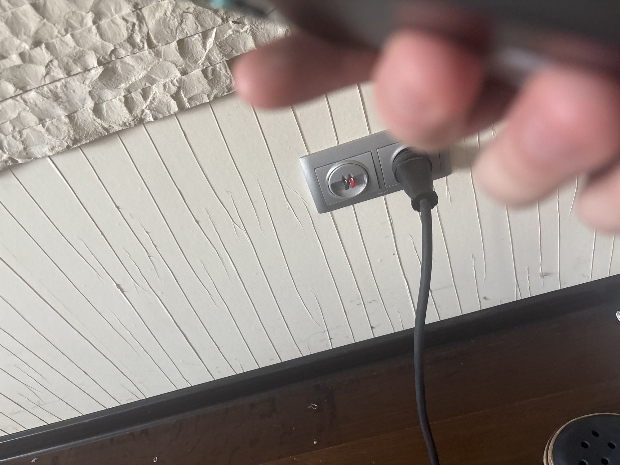 The image shows a close-up view of a wall with horizontal white panels. To the left, a dark brown wooden surface is visible, possibly a door frame or furniture. A black electrical cord is plugged into a silver electrical outlet on the wall. The outlet has two rectangular sockets and a circular socket above them, which appears to have a red light indicating power. The wall has a textured surface with visible creases or folds in the panels. In the foreground, out of focus, are parts of a person's hands and fingers, with one finger appearing to be near the electrical outlet. The lighting suggests an indoor setting, possibly a residential or commercial space. There is no information about the location in the city of Ореховка, Russia.