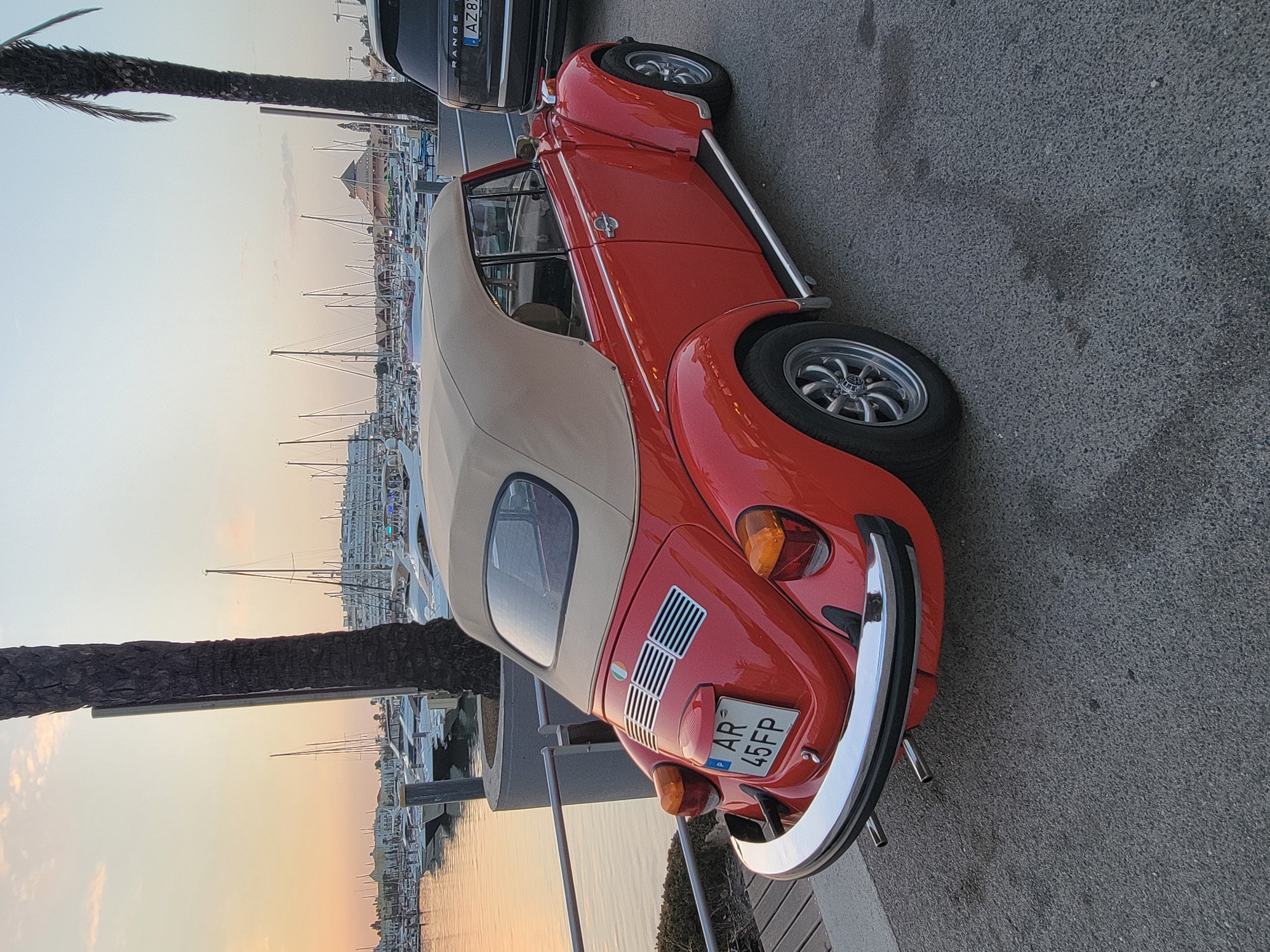 A bright red, classic Volkswagen Beetle convertible with a tan soft top is parked on an asphalt surface next to a marina in Quarteira, Portugal. The car's license plate reads "AR 45FP". In the background, numerous sailboats and larger yachts are docked in the water, with buildings and a domed structure visible on the distant shore. A dark-colored SUV is parked behind the Beetle. The sky is a soft gradient of orange and yellow, indicative of sunset or sunrise. Palm trees are present in the foreground and to the right, with one on the left partially obscuring the view. A metal railing and a concrete barrier separate the parking area from the marina. The lighting suggests it is either early morning or late evening.