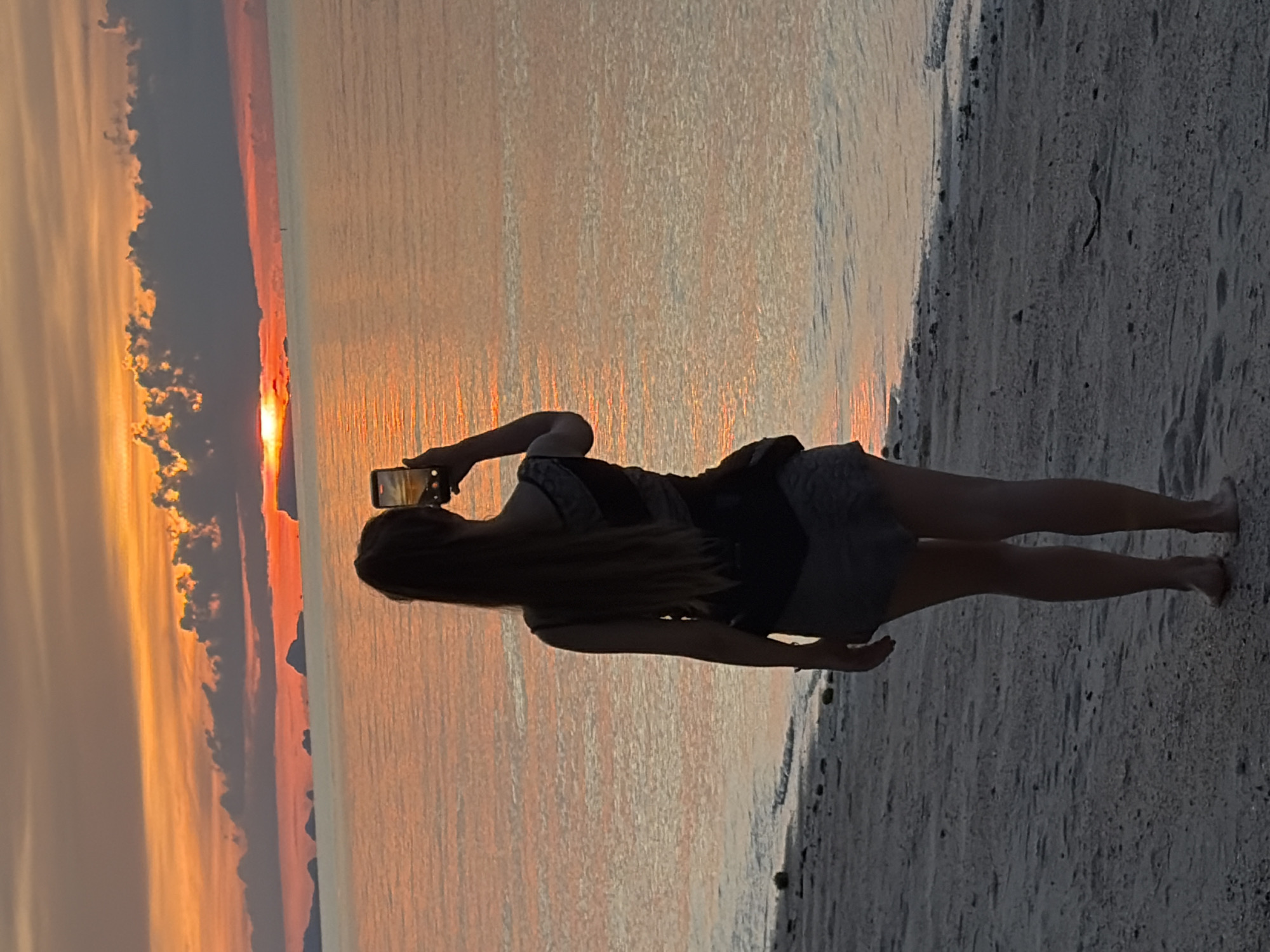 A woman stands on a sandy beach in Baan Tai, Thailand, facing the ocean and holding a smartphone up to take a picture of the sunset. The sky is filled with vibrant orange and pink hues, with dramatic dark clouds partially obscuring the setting sun. The ocean reflects the colorful sky, creating a shimmering effect. In the distance, several islands are visible as silhouetted shapes against the horizon. The woman is wearing a dark top and light-colored shorts, with her long hair flowing down her back. The sand on the beach is light-colored and appears wet near the water's edge, with small pebbles and debris scattered across it. The overall scene captures a serene and picturesque moment at sunset.
