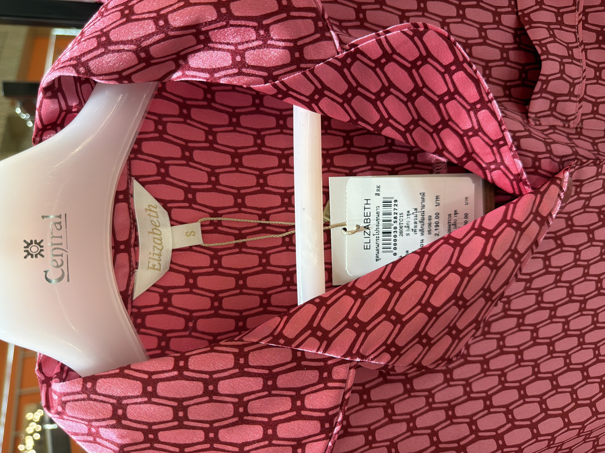 The image shows a close-up of a pink patterned shirt hanging on a white hanger in a retail environment. The shirt has a repeating geometric pattern of interconnected hexagonal shapes in a darker pink. A white tag is attached to the collar, displaying the brand name "Elizabeth" in gold lettering. Below "Elizabeth" on the tag, there is text in Thai script and a size "S". Another white tag, partially visible, contains product information in Thai script and numbers, including a price. The background is blurred but suggests an indoor setting, possibly a store, with bright lighting. The location is identified as Ko Samui District, Thailand.