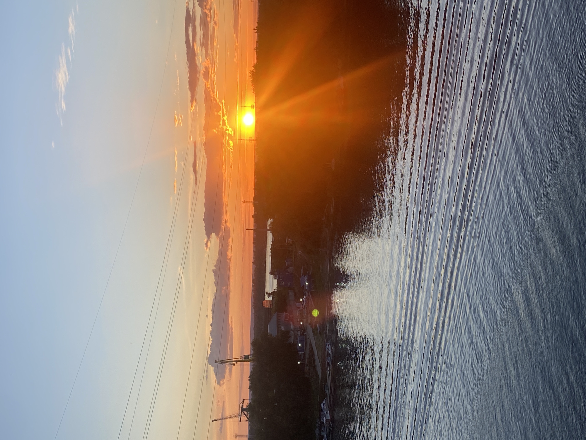 The image captures a sunset over the Volga River in Novokuybyshevsk, Russia. The sun, a bright orange disc, is low on the horizon, casting a warm glow across the sky and water. Its rays fan out, creating a dramatic effect. The river's surface is rippled, reflecting the colors of the sky. Along the far bank, there is a line of trees, and further in the distance, several buildings are visible, including what appear to be small houses or industrial structures. Cranes can be seen near the shore, suggesting a port or industrial area. Power lines crisscross the sky, adding a modern element to the natural landscape. The overall scene is one of natural beauty enhanced by the industrial backdrop.