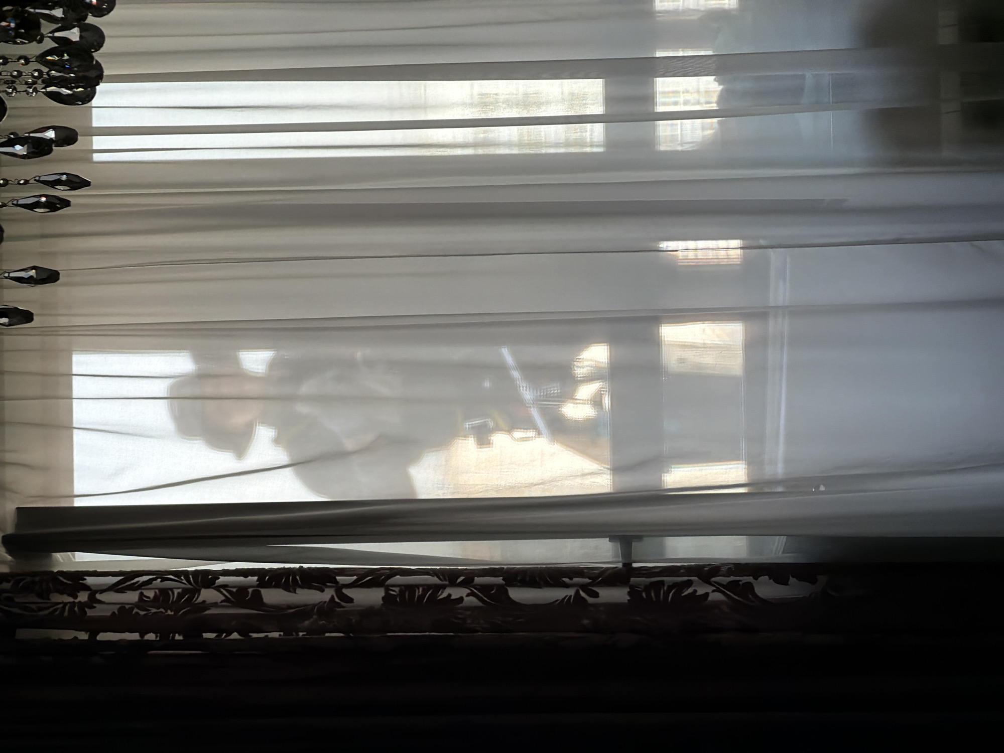 The image shows a window covered by a light-colored, sheer blind or curtain with horizontal slats. There is a beaded curtain trim hanging to the left of the window. Outside, a blurry reflection of a person and furniture is visible through the blinds. The lower portion of the window is obscured by a patterned valance, and below that, dark furniture is visible. The blinds or curtain appear to be inside a room in Khimki, Russia.