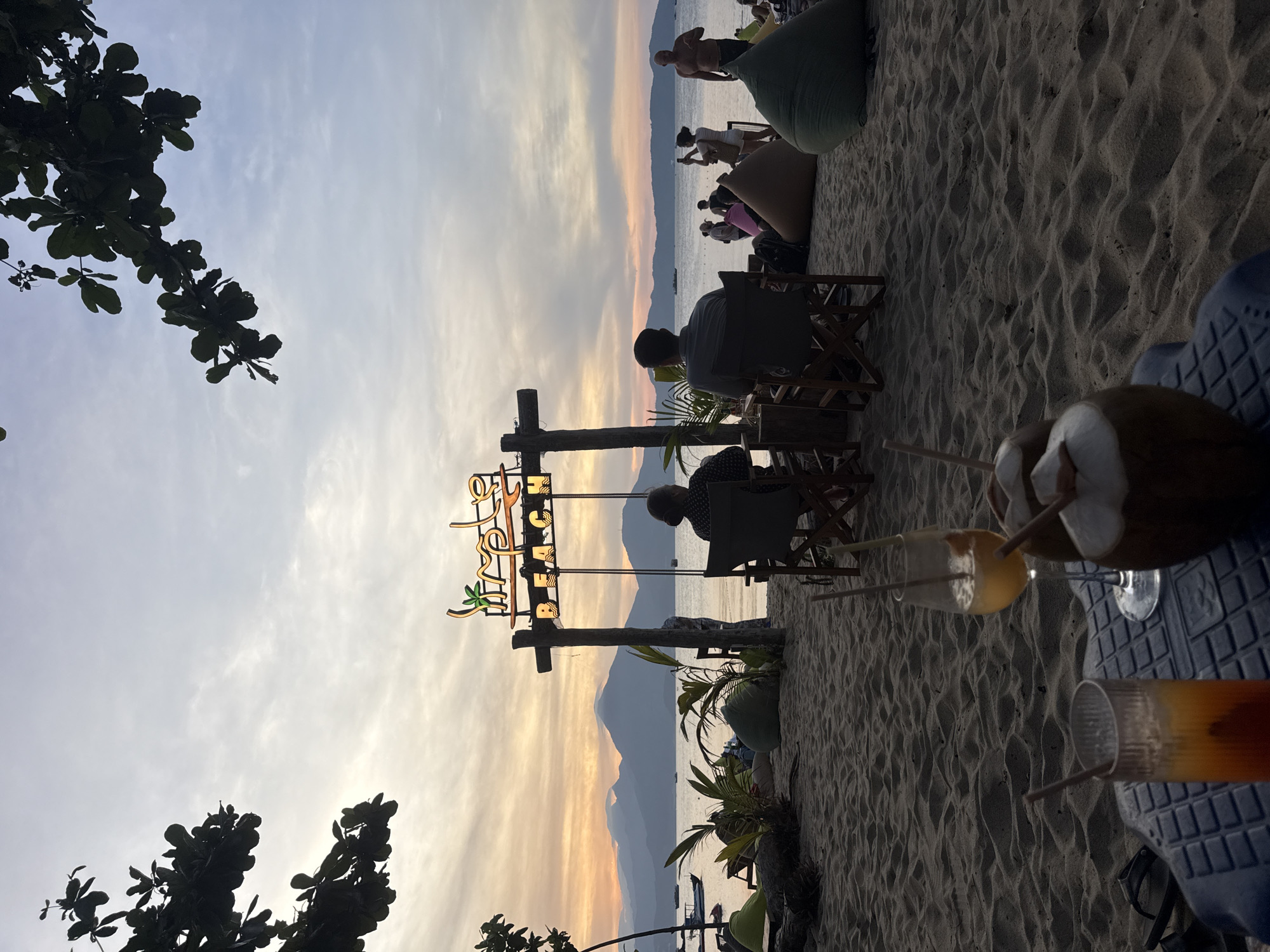 The image depicts a beach scene at sunset in an unknown city. Several individuals are visible, including people seated at wooden tables and chairs, others relaxing on beanbag chairs, and some standing near the shoreline. The sky displays a gradient of orange and grey hues, reflecting off the ocean. In the foreground are drinks, including a coconut with a straw. A sign with the words "The Beach Club" is erected on wooden posts. The scene is framed by foliage on the left side and sandy beach details on the right. A mountain range is visible in the distance.