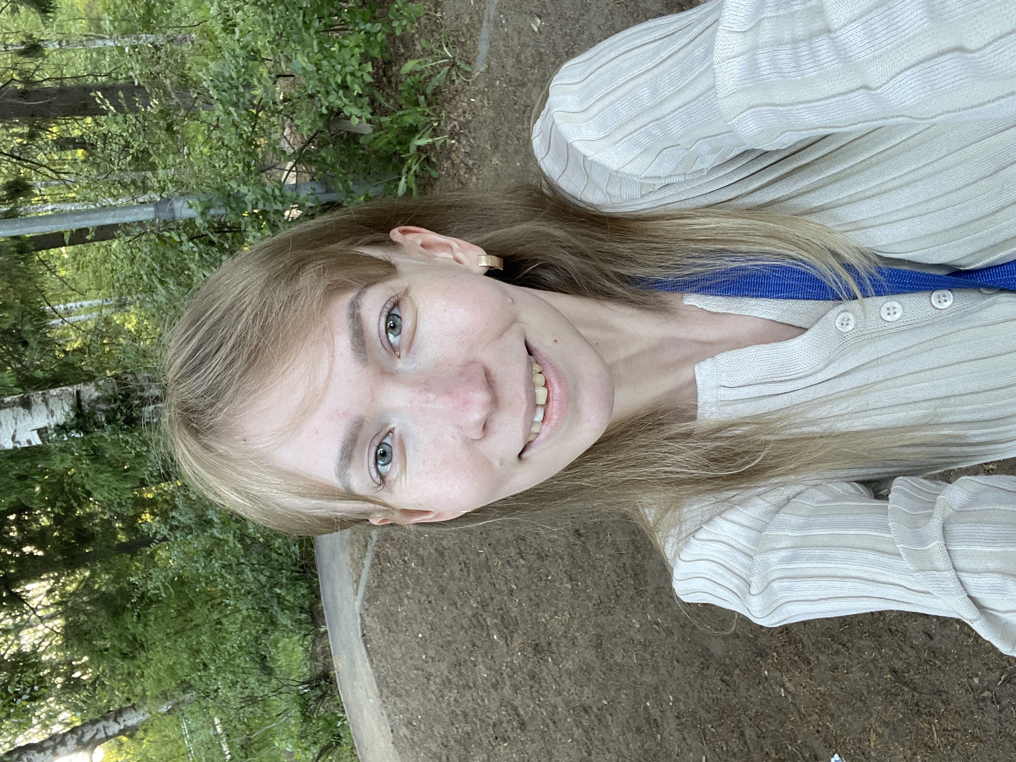 A medium close-up shot shows a woman with blonde hair, pale skin, and green eyes smiling slightly at the camera. She wears a cream-colored, ribbed cardigan over a blue top. An ear with a gold earring is visible.
The background suggests an outdoor location, with green foliage and trees visible at the top and left of the frame. The ground appears to be dirt or mulch. The city of Perm, Russia, is not discernible from the image.