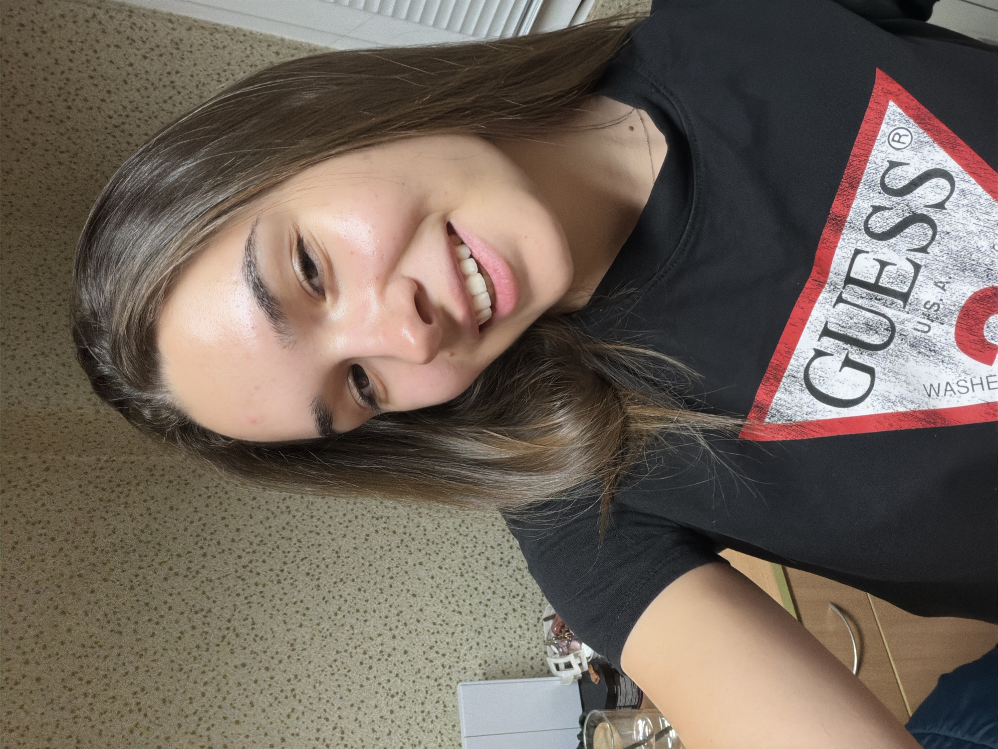 A young woman with long, brown hair is smiling and looking at the camera while wearing a black Guess t-shirt. The background includes what appears to be a window with blinds, a speckled surface resembling a countertop, and a wooden floor. The location in Sofia, Bulgaria cannot be determined based on the given information.