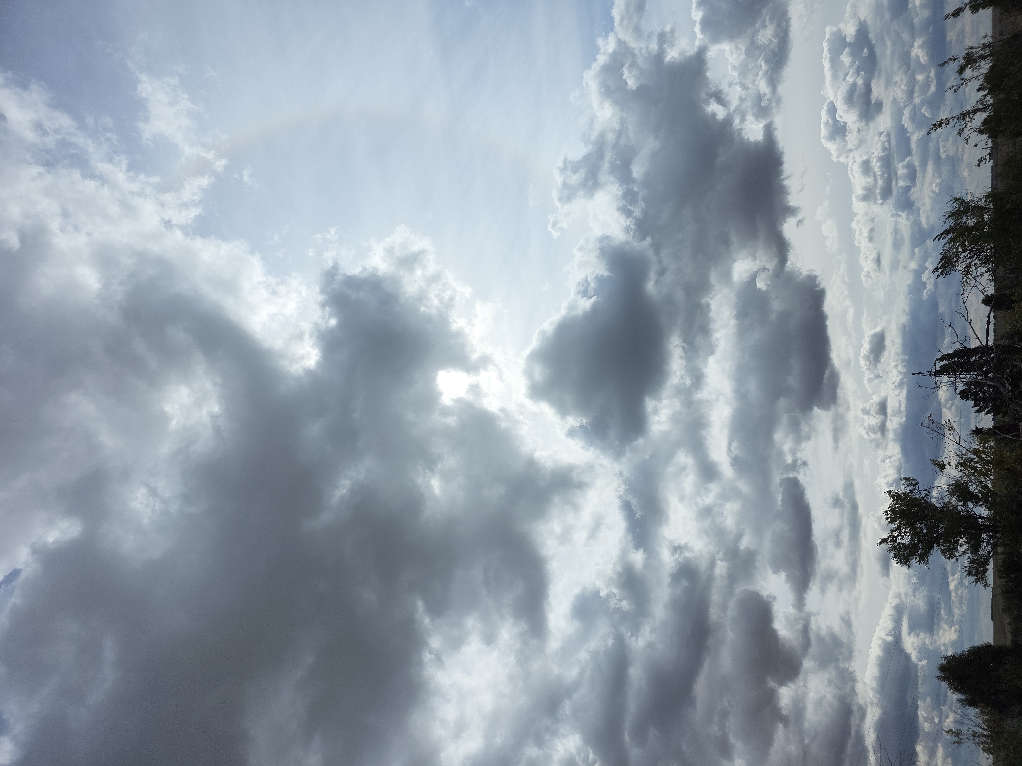 The image captures a dramatic sky over Qarabalıq, Kazakhstan, characterized by a thick layer of dark, turbulent clouds dominating the upper frame. Sunlight struggles to break through, creating a diffused glow in some areas and casting shadows on others. A faint arc, possibly a sun halo or a weak rainbow, is visible in the upper right portion of the sky, hinting at the interaction between sunlight and moisture in the atmosphere. Below the dense cloud cover, a horizon of silhouetted trees and vegetation is discernible. The landscape appears somewhat sparse, suggesting a natural or semi-rural setting. The overall mood is one of impending weather change or the aftermath of a storm. There are no people or discernible actions taking place within the frame; the focus is solely on the atmospheric conditions and the natural landscape.