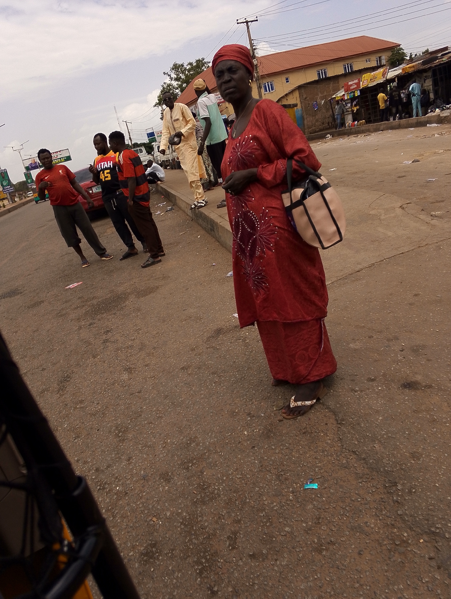 The image depicts a street scene in Jos, Nigeria. In the foreground, an older woman wearing a red dress, headwrap, and flip-flops stands on the roadside carrying a cream-colored tote bag. Behind her, a group of men are gathered. One man wears a red t-shirt, another a basketball jersey with the number 45 and the word "UTAH". Further back, there is another man in light-colored traditional attire. A red car is parked near the group. The street is littered with debris. In the background, buildings and commercial stalls are visible, with people milling about. Overhead power lines are present. A portion of a vehicle is in the lower left corner of the image, obscuring the view.