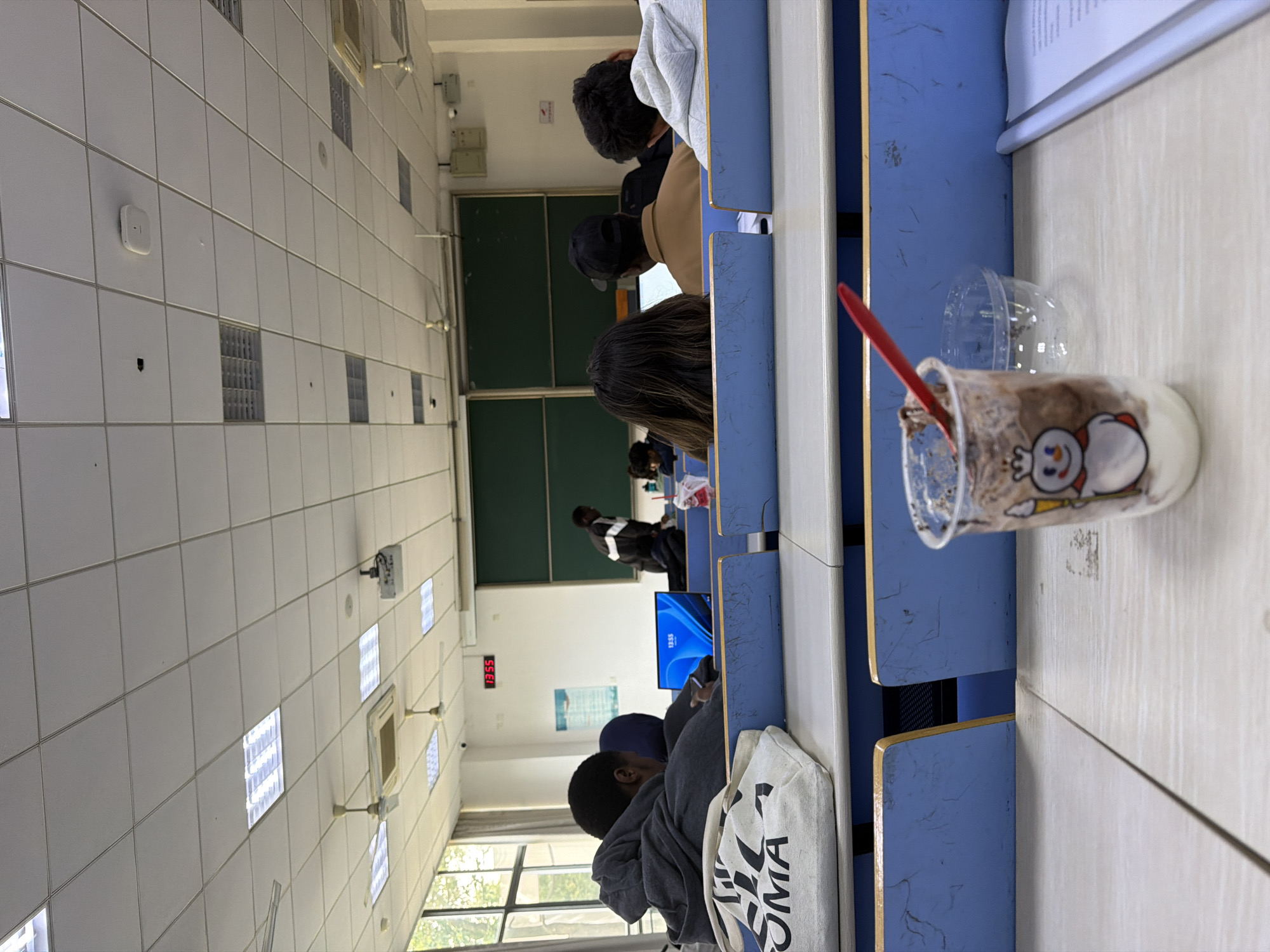 The image shows a classroom setting in what appears to be a university or college in Jiangning District, China. Students are seated at desks, focused on individual laptops. The desks are blue and white. A plastic cup with an image on it and a red spoon inside is on the desk in the foreground. The room is lit by fluorescent lights, and there is a digital clock on the wall. A whiteboard is also visible.