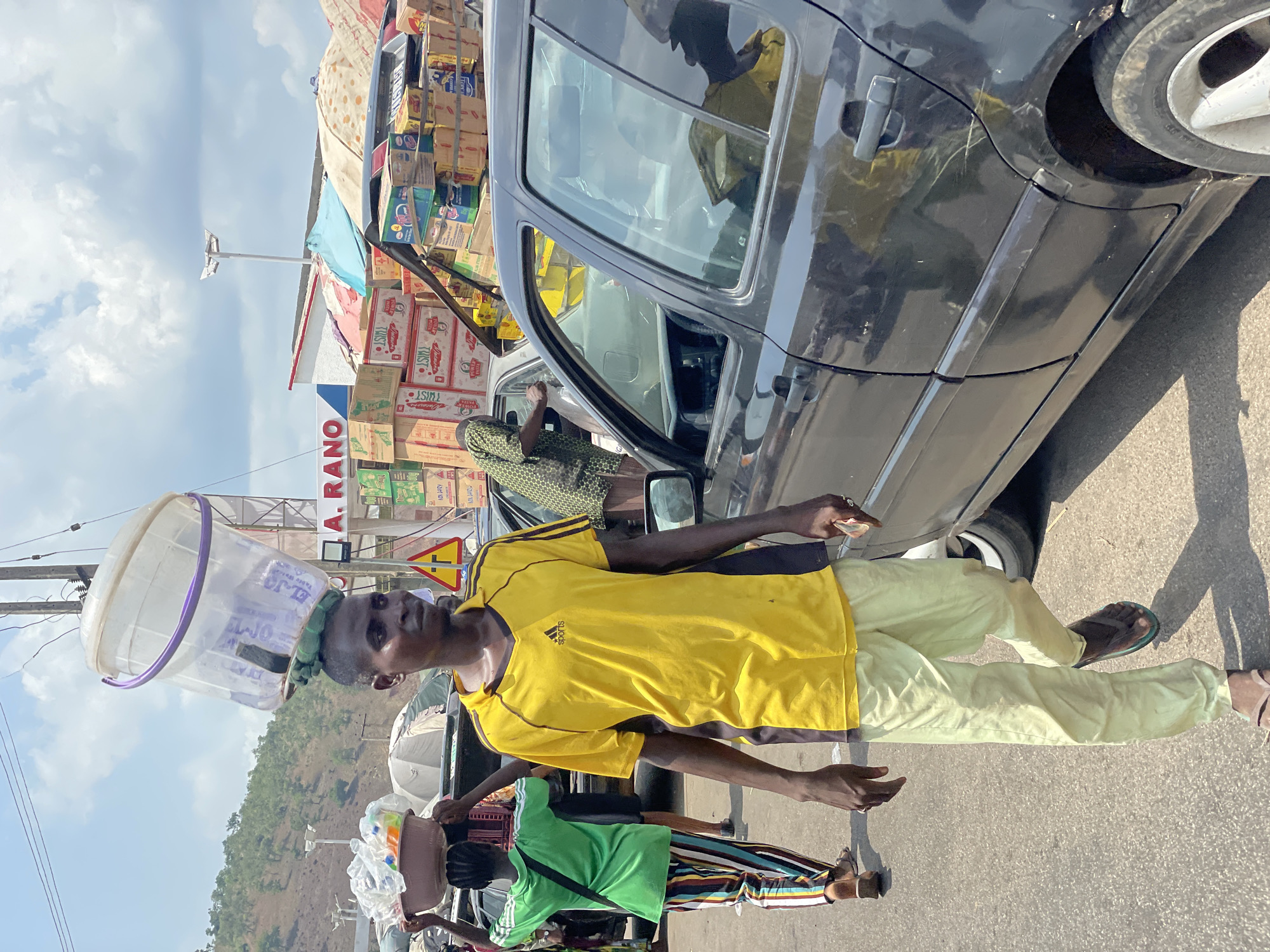 In Lokoja, Nigeria, an individual in a yellow Adidas shirt and light green pants stands on a paved road next to a parked black car. They have a purple-rimmed plastic bucket balanced on their head. Another person in a green shirt and striped pants walks beside them, also carrying an object on their head. In the background, there are stacks of boxes with the name "A. RANO" visible. A mountain is also visible.
