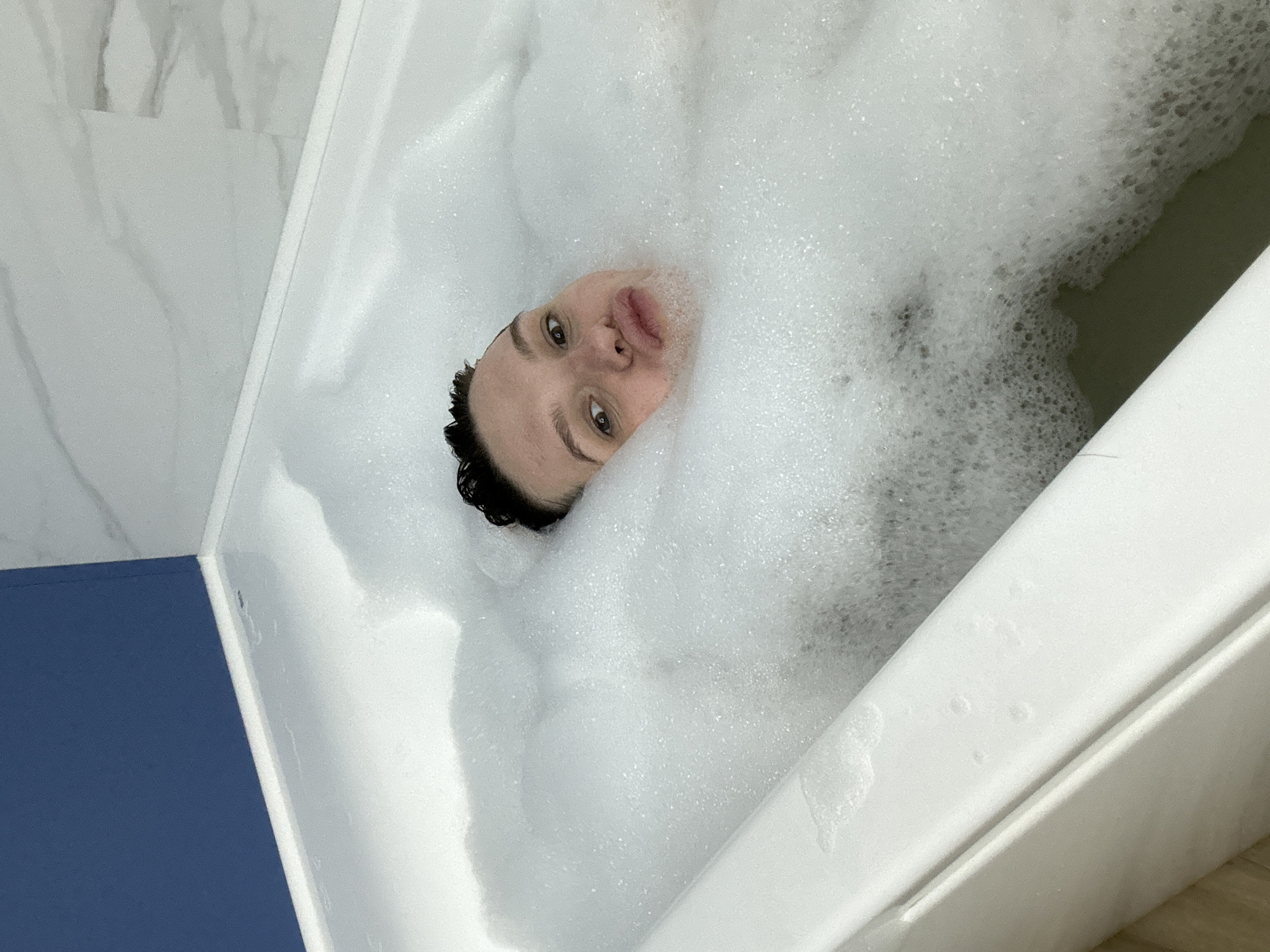 A woman with short, dark hair is submerged in a bathtub filled with white bubbles. Her face is visible just above the bubble line. The bathtub is white and has a blue panel on the side. The location is inferred to be in the city of Chaikovsky, Russia. The photograph's composition is taken from a high angle, partially rotated clockwise.