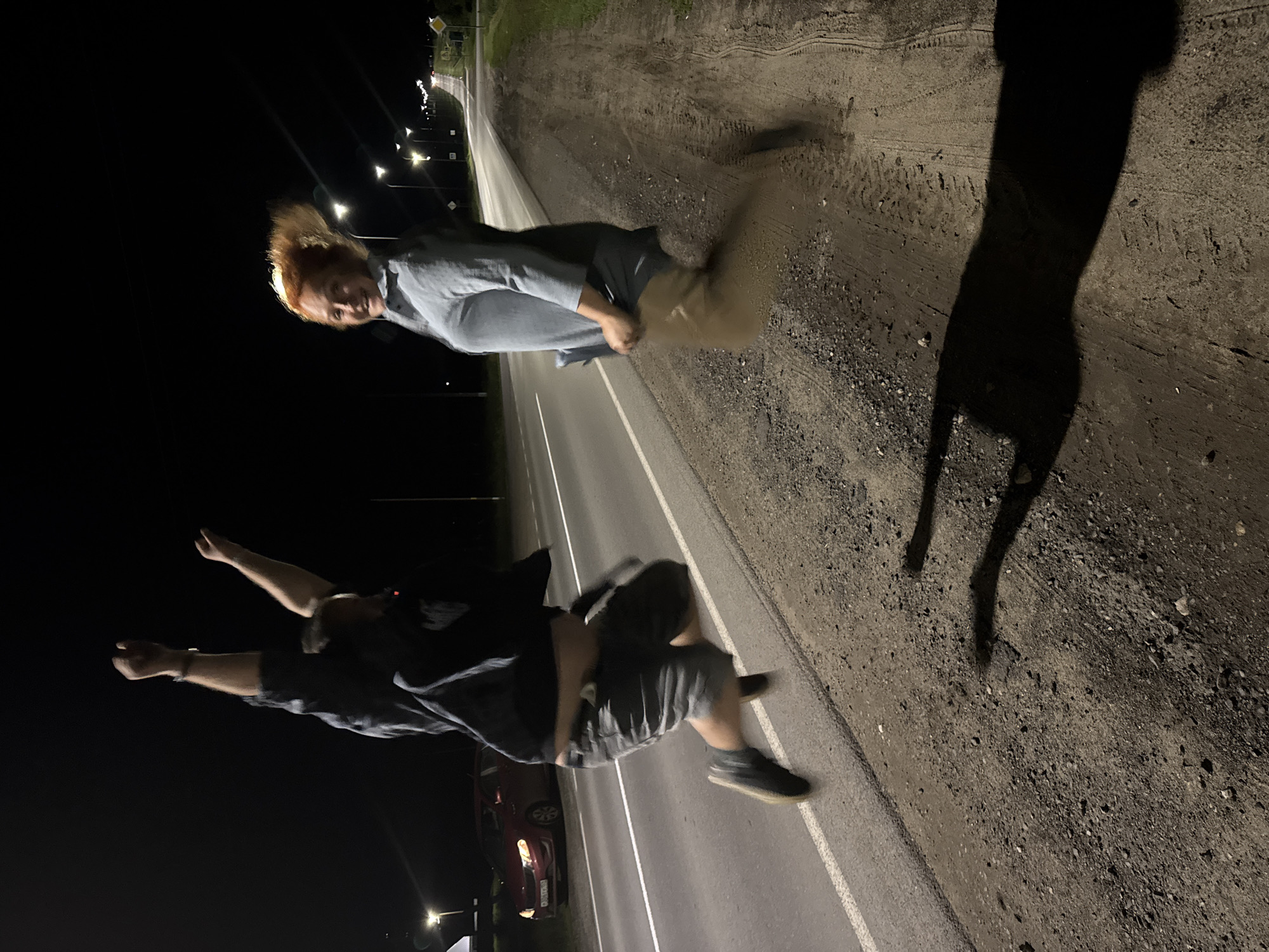 Two individuals are captured mid-air, jumping on the shoulder of a paved road at night. The person on the left, with dark hair, is wearing a dark t-shirt and grey shorts, with their arms raised in a celebratory gesture. The person on the right, with red hair, is wearing a light blue long-sleeved shirt and appears to be looking towards the camera with a smile. The road is illuminated by streetlights in the background, and a red car is parked on the side of the road. The ground consists of dirt and gravel. The shadows of the jumping individuals are cast on the ground. The location is identified as Anatazh, Russia.