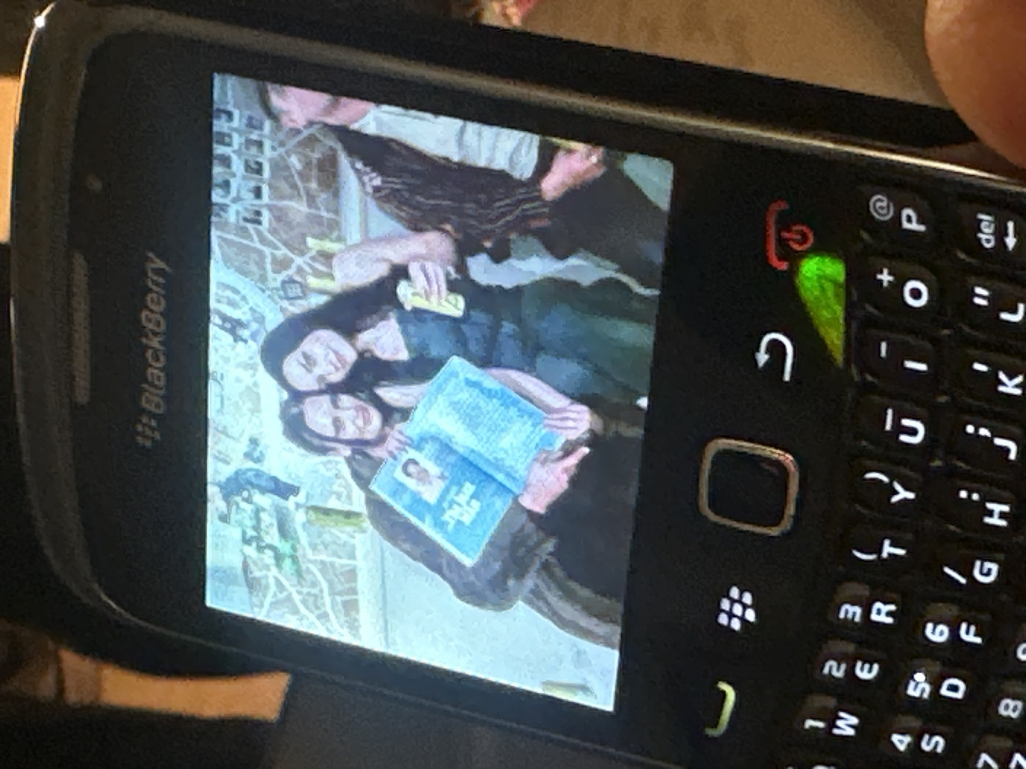 A close-up image shows a black BlackBerry smartphone held by a human hand, displaying another image on its screen. The phone features a full QWERTY keyboard, with illuminated keys, a green indicator light near the return key, and a red power symbol. The screen displays a digital photograph featuring three individuals positioned in front of a textured wall adorned with numerous small, square photographs or polaroids.
From left to right within the displayed image:
1. A person with dark hair, wearing a dark long-sleeved garment, holds an open blue booklet or magazine. A small photo of a man and text, possibly "THE NEW MAN," are visible on its left page. The person's left hand points towards the booklet's lower section.
2. A woman with long dark hair, wearing a dark top, smiles directly towards the camera while holding a light-colored bottle or can in her right hand.
3. A male figure is partially visible on the right, wearing a patterned vest over a light-colored shirt.
The background consists of a light-colored, possibly concrete or brick wall, covered in a collage of small, square photographs. Some green elements, possibly foliage or graffiti, are also visible. The overall environment, characterized by street art and public photo displays, is indicative of urban art districts such as Shoreditch or Brick Lane in the city of London, United Kingdom.