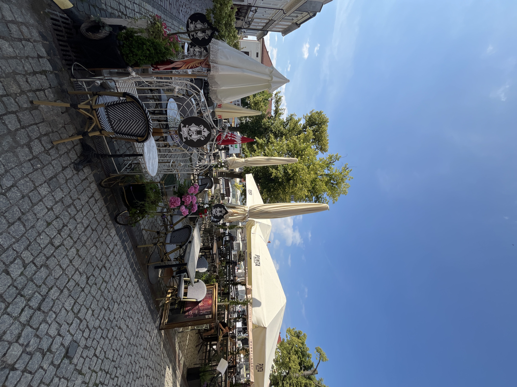 The image depicts an outdoor seating area of a cafe on a cobblestone street. Several tables and chairs are arranged under large umbrellas, some of which display the brand "PINTA". Decorative elements like white wrought-iron furniture, potted plants, and signage are visible. In the background, trees and buildings line the street. The sky is clear and blue with a few wispy clouds. One person is seated at a table in the mid-ground, facing away from the camera. The scene appears to be set during daytime, with sunlight casting shadows on the ground.