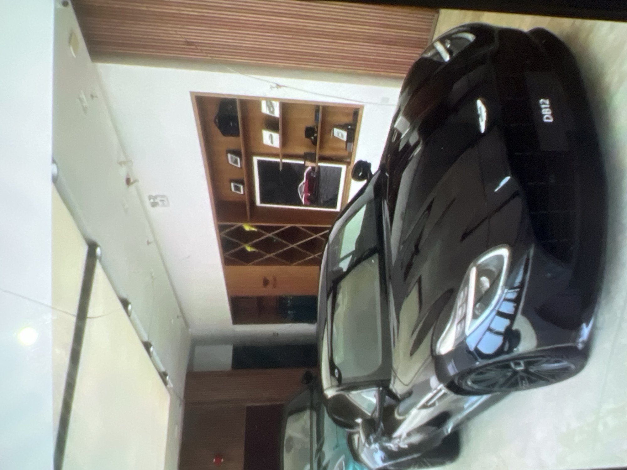The image shows a black Aston Martin DB12 parked in what appears to be a showroom. The car is the primary subject, with its sleek design and headlights prominently visible. In the background, another car, possibly teal, is partially visible. The showroom features wooden paneling on the walls and a shelving unit displaying framed pictures and objects. The lighting in the room is soft, with ceiling lights providing illumination. There is no indication of the location being in the city of Kara-Dıykan, Kyrgyzstan, or any other specific geographical location. The scene is static, with no people or actions depicted.