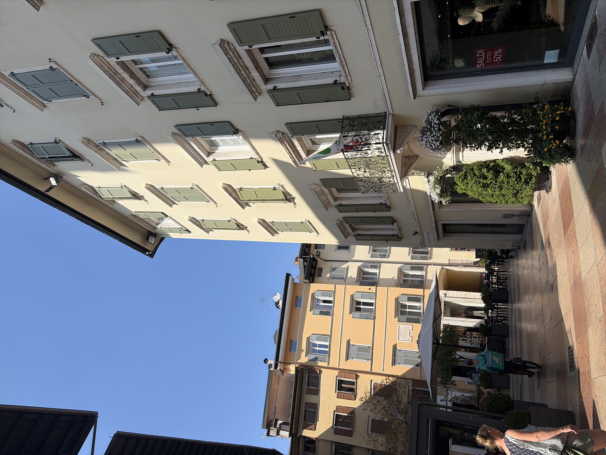 A bright, sunny day illuminates a pedestrianized street in Trento, Italy, lined with historic buildings. The architecture features cream and yellow facades with traditional green shutters, many of which are partially open. A narrow street opens up to reveal more buildings and a clear blue sky. In the foreground, a woman walks away from the camera, carrying a green bag. Further down the street, a delivery cyclist with a teal backpack rides towards the viewer. The ground is paved with patterned stones. A large, grey umbrella shades an outdoor seating area in front of a building. The right side of the image shows a shopfront with a display window featuring a mannequin and a "SALDI TUTTO -50%" sign. The overall impression is of a vibrant and charming Italian urban environment.