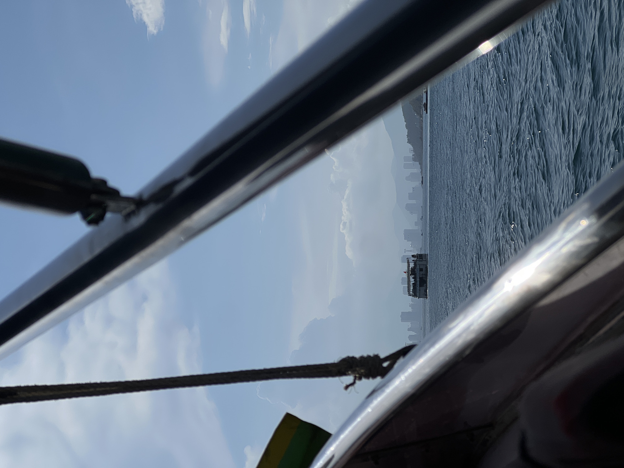 The image shows a view from a boat, with metallic railings and a rope in the foreground. The background features a body of water with ripples, possibly the ocean, and a distant city skyline under a partly cloudy sky. A larger boat or ship is visible on the water in the middle distance. The location is Nha Trang Ward, Vietnam.