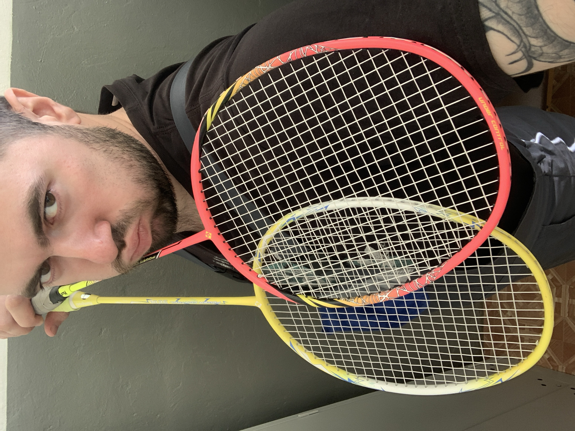 A man with a beard and dark hair is holding two badminton rackets. The top racket is pink and orange. The bottom racket is yellow and white. The man is wearing a black shirt. Part of an arm with a tattoo is visible. The location is unknown.