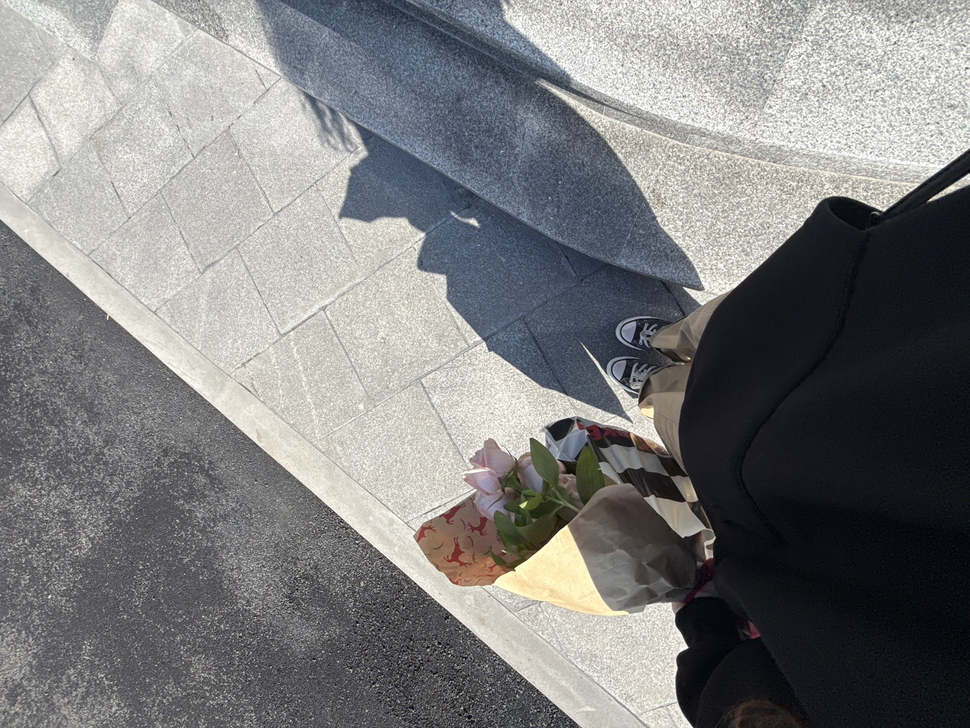 The image shows an individual standing on a grey brick sidewalk next to a grey ledge, possibly a step. The individual is wearing black sneakers, light tan pants, and a black jacket. They are holding a bouquet of light-colored roses wrapped in brown paper. The shadow of a person is cast on the sidewalk. The image may be taken in Yerevan, Armenia, but that can't be verified from the image alone.
