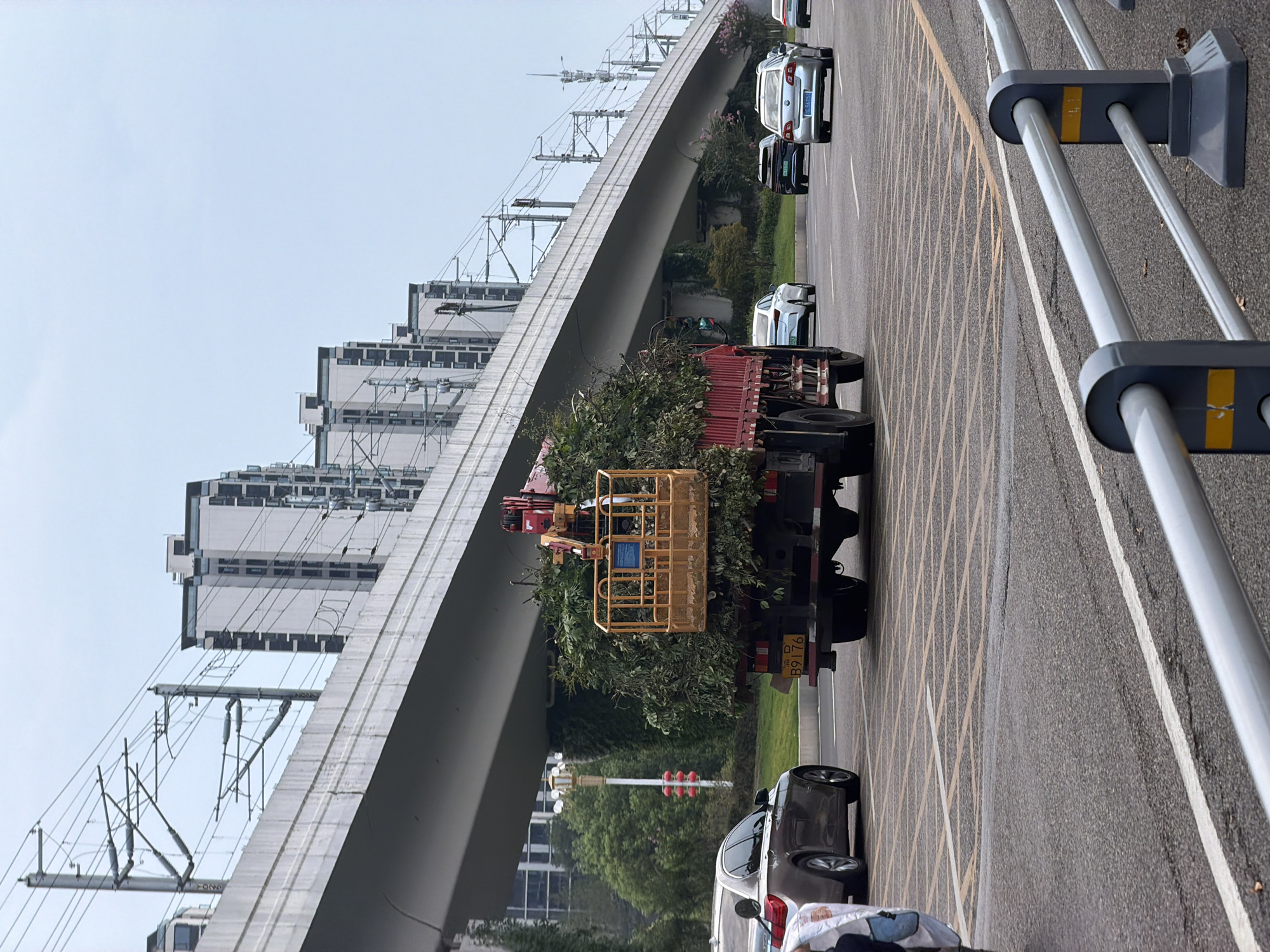 The image shows a street scene in Jiangjin, China, with a truck carrying a large load of branches and foliage. The truck has a yellow basket lift attached to its side, suggesting it may have been used for pruning or tree removal. Several cars are visible on the road, including a silver sedan and a white sedan. In the background, a multi-story elevated highway or bridge structure is prominent, with power lines and pylons running parallel to it. Beyond the bridge, several modern buildings with multiple stories are visible. The sky is overcast. A traffic light with red lights illuminated is visible in the distance. A metal barrier with yellow markings is in the foreground. The scene appears to be a typical urban environment with ongoing traffic and infrastructure.