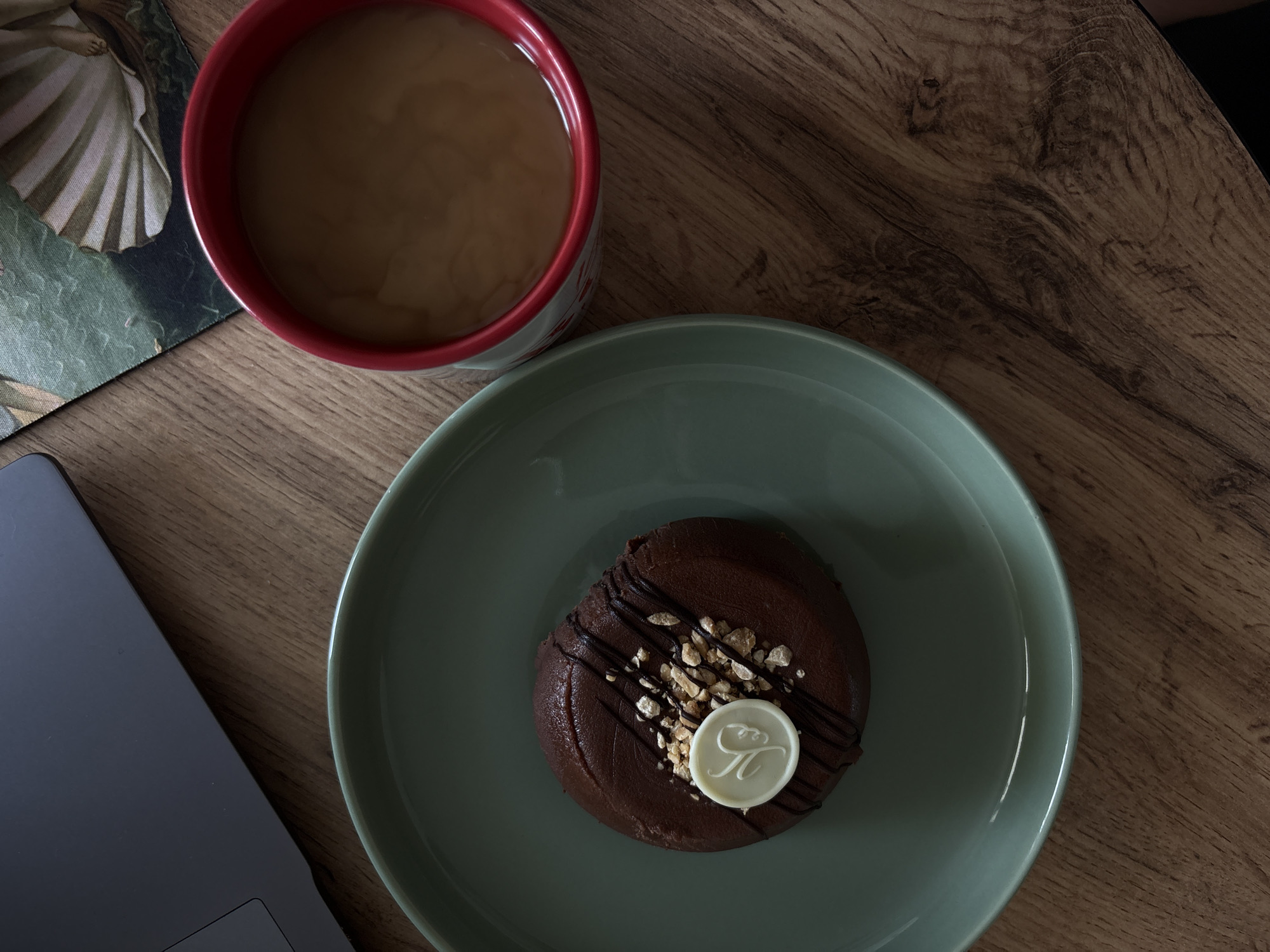 A top-down view shows a chocolate cake on a teal plate placed on a wooden table. The cake is topped with chopped nuts, a drizzle of dark chocolate, and a small white chocolate disk with a decorative imprint. To the right of the plate is a red mug filled with a light brown beverage, possibly coffee with cream. In the background, a portion of a laptop is visible on the left, and a postcard or artwork with a green and white design is on the right. The scene is set on a wooden surface that appears to be a table. There are no people visible in the image, and no specific details suggest a location in Rostov-on-Don, Russia.