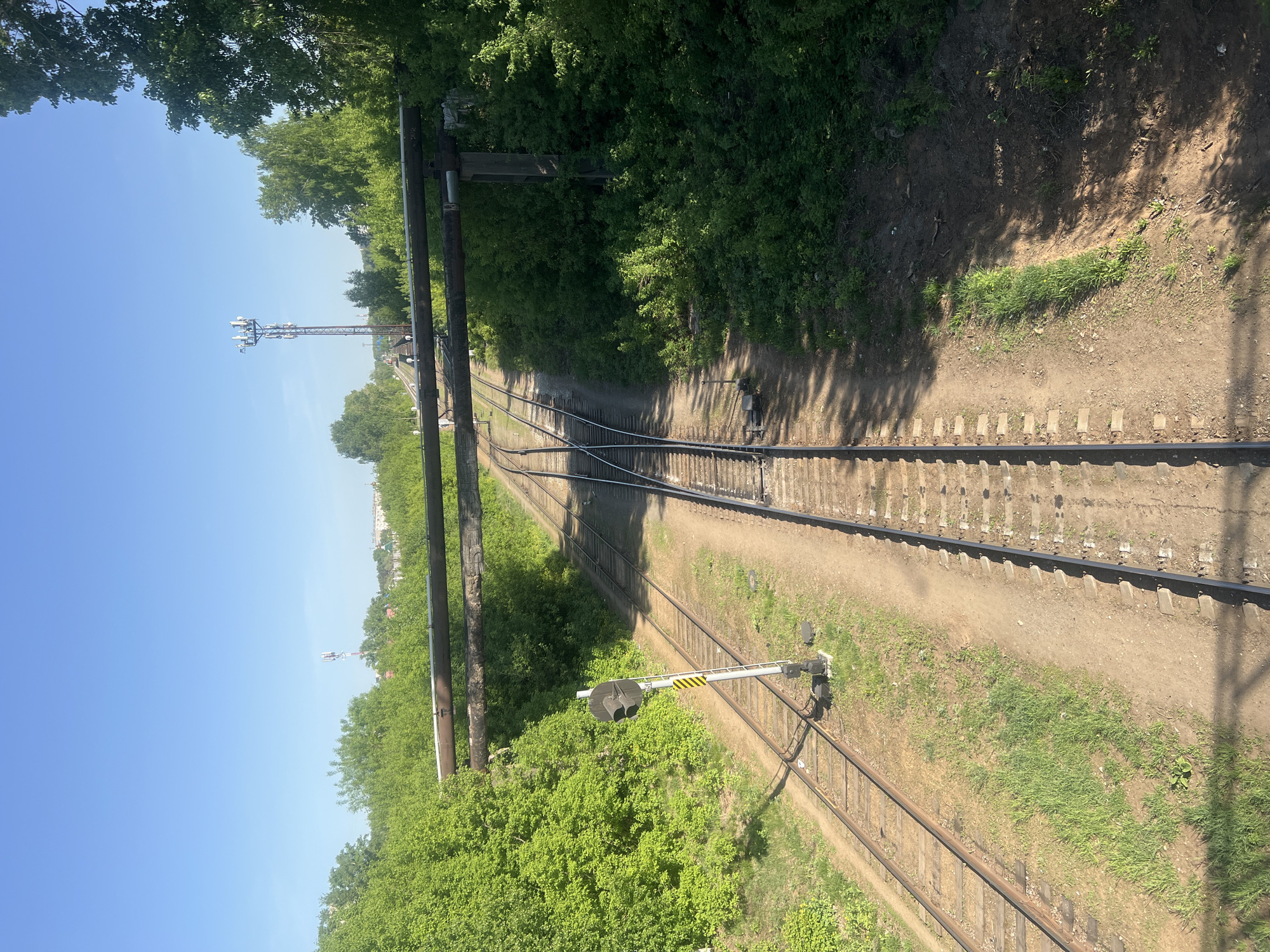 An elevated, downward-facing shot shows two parallel railroad tracks converging in the distance. A rail signal post sits adjacent to the right track, while a set of parallel pipes extend horizontally across the left side of the frame. Green vegetation lines both sides of the tracks, suggesting a somewhat rural or undeveloped area. A tall communications tower is visible in the distance, hinting at a possible urban or suburban location. The clear blue sky suggests daylight.
The scene is in the city of Chaikovsky, Russia.