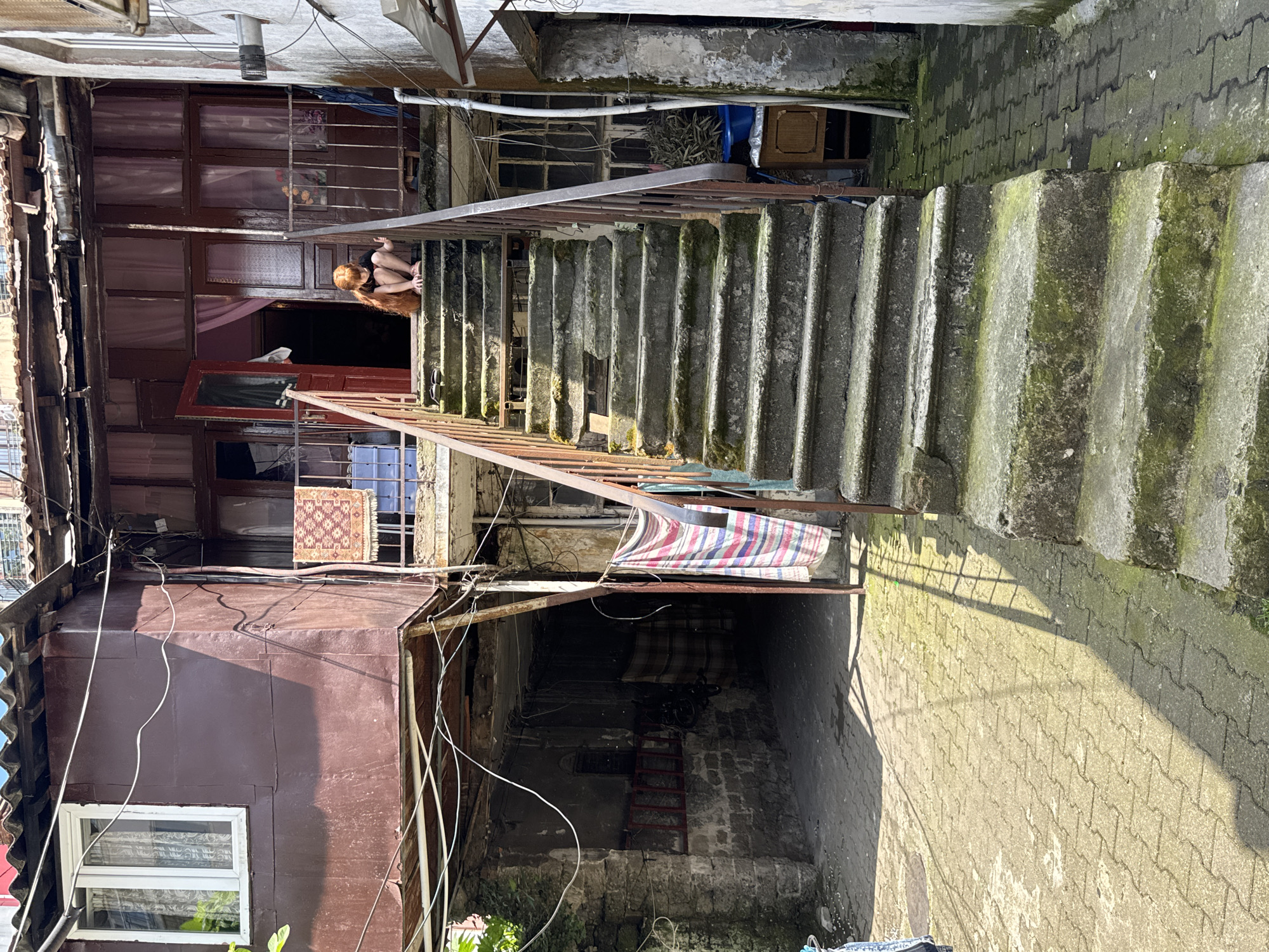 The image depicts a narrow alleyway in what appears to be a densely populated urban area, possibly Batumi, Georgia. The alleyway is flanked by dilapidated buildings on either side, constructed from a mix of materials including corrugated metal, brick, and concrete. An external concrete staircase dominates the center of the image, ascending from the bottom to the upper level. The stairs are partially covered in moss.
A person with long reddish-brown hair is seated on the top landing, partially obscured by the staircase railing. The buildings show signs of disrepair, with peeling paint, exposed wiring, and weathered surfaces. Various items are hanging from the buildings, including rugs and cloth, adding to the cluttered appearance. The ground is paved with interlocking concrete blocks. In the lower section of the alleyway, a red ladder leans against a brick wall. A bicycle is visible in the background.