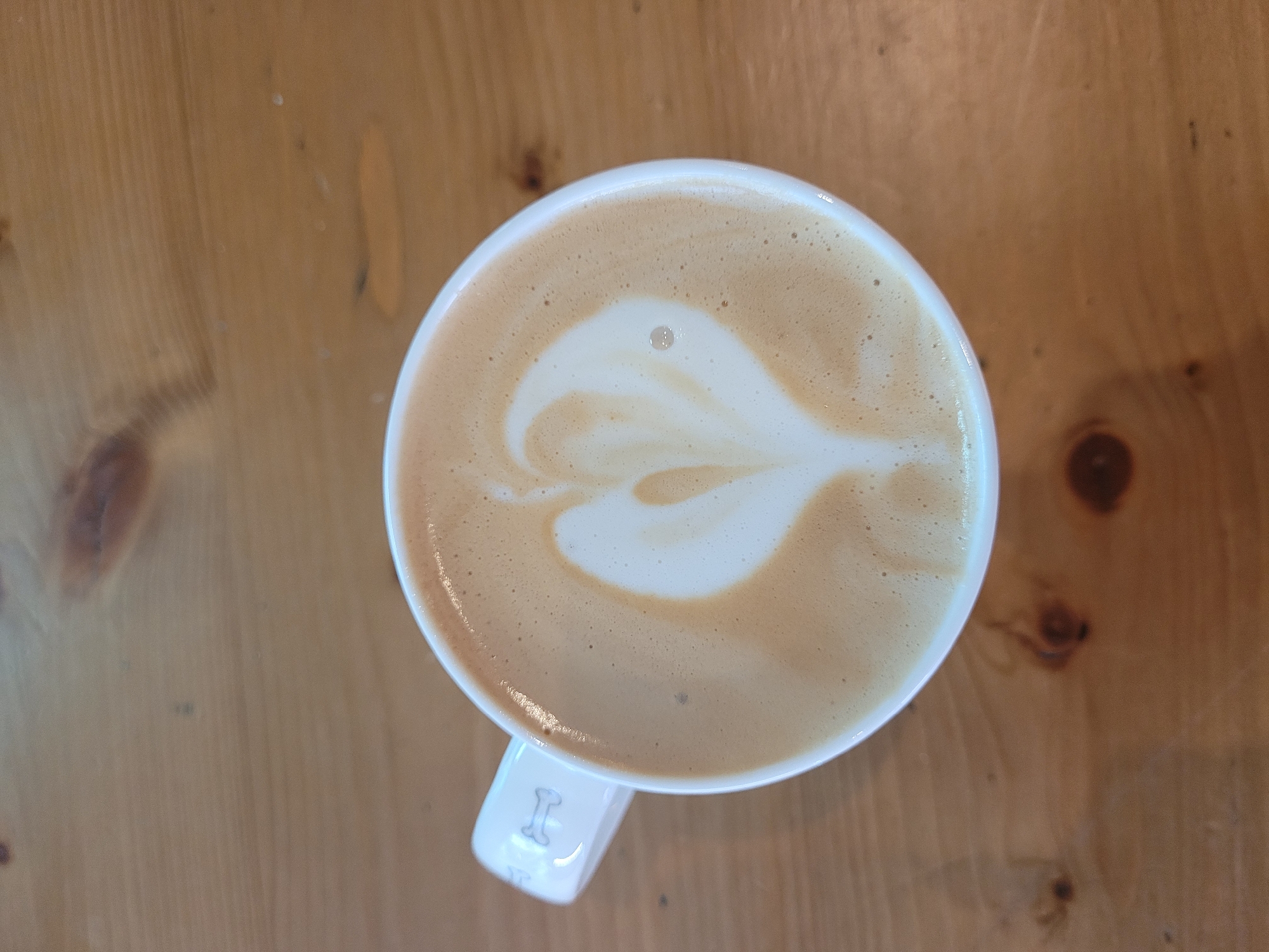 An overhead, medium shot shows a white ceramic mug of latte art with a heart design, placed on a light brown wooden table. The mug has a white handle with a subtle grey dog bone design. The latte art is composed of white milk foam against the brown coffee. The table has visible wood grain and knots. The background is out of focus. The scene is indoors. This image was taken in Limassol, Cyprus.