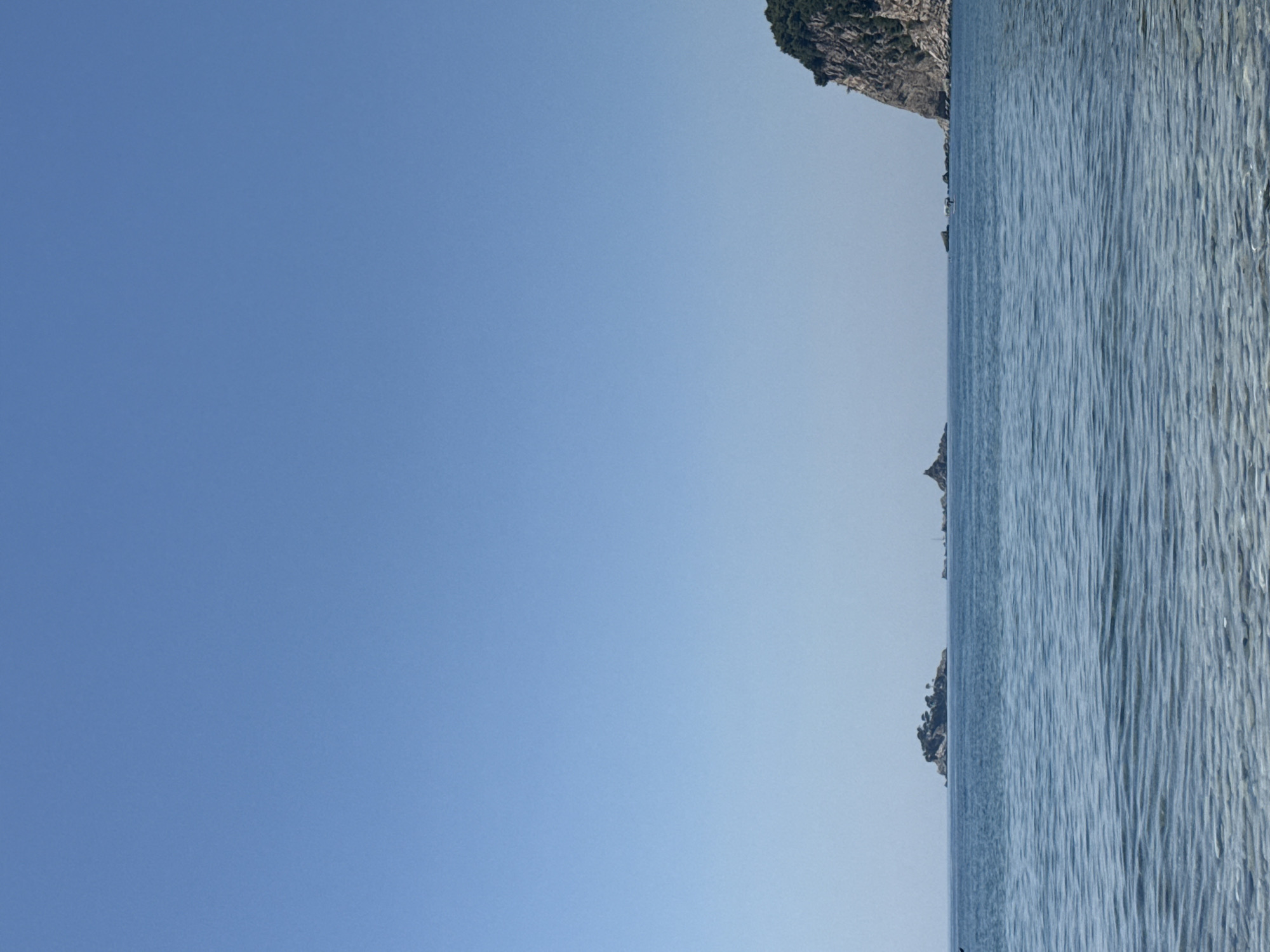 The image depicts a coastal scene in Buljarica, Montenegro, characterized by a clear blue sky and calm, blue waters. In the foreground, the shallow water near the shore reveals a sandy seabed with a subtle ripple pattern on the surface. The middle ground is dominated by the vast expanse of the sea, with a few small, rocky islets scattered across it. One prominent island, covered in sparse vegetation, is visible to the left. Further to the right, a larger, rocky cliff face covered in lush green trees forms the backdrop on the right side of the frame. The overall impression is one of a tranquil, sunny day by the Adriatic Sea.