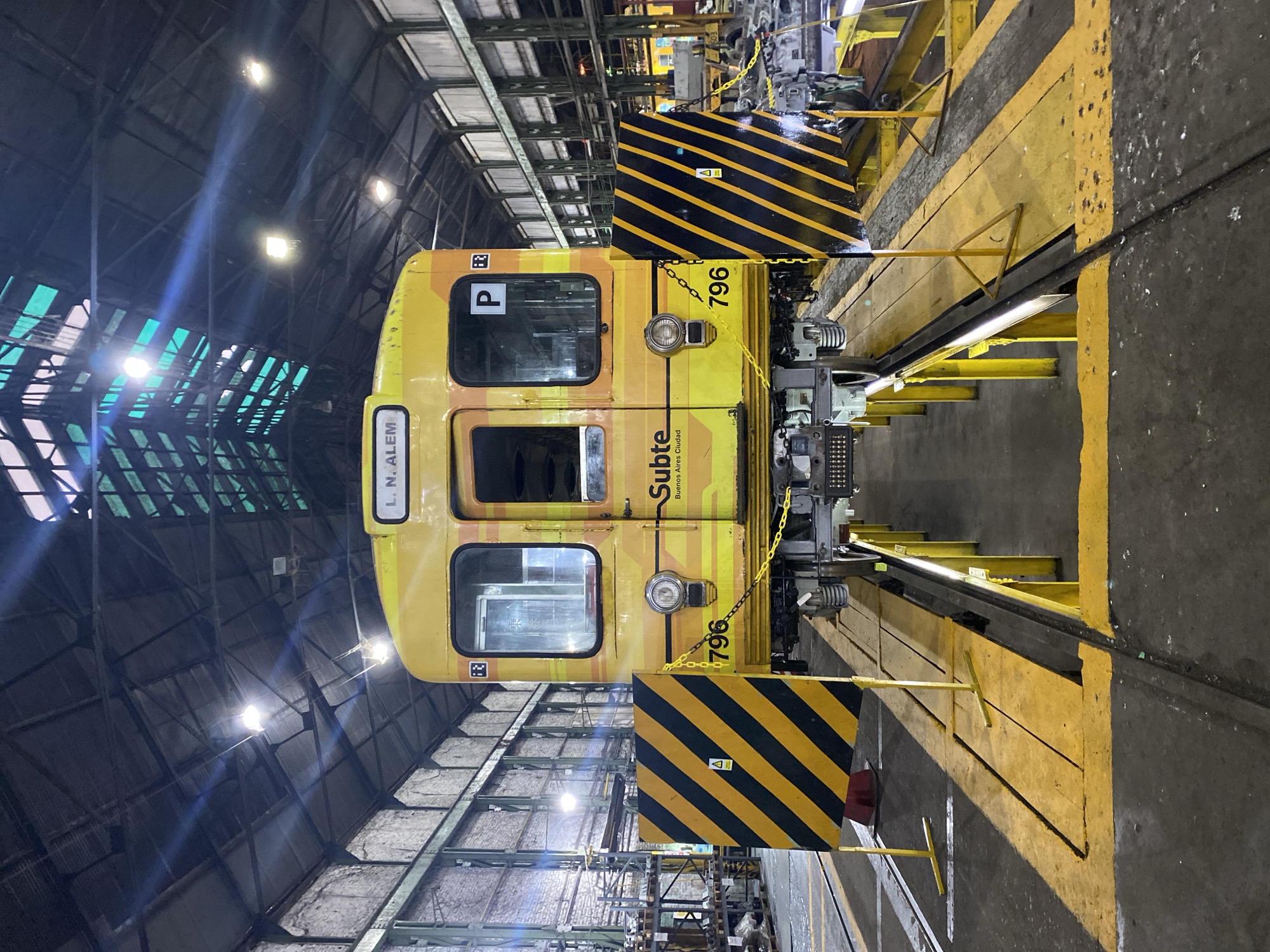 The image displays a yellow and orange Mitsubishi 5000 Series subway car, designated with the number "796" on its lower front, situated within the Taller Lacroze (Lacroze Workshop), a Subte maintenance depot located near the Federico Lacroze station in the Chacarita neighborhood of Buenos Aires, Argentina. The train car, part of the Buenos Aires Subte system (indicated by "Subte Buenos Aires Ciudad" on its side), exhibits "L.N. ALE" on its front destination display, signaling Leandro N. Alem as a destination. A black letter "P" is visible on a white background within a side window of the car.
The subway car is elevated on yellow metal support structures above a maintenance pit, exposing its undercarriage and wheel assemblies. The pit opening is partially covered by wooden planks, and the surrounding concrete floor is marked with yellow safety lines. Two black and yellow striped safety barriers, each with yellow support legs, are positioned directly in front of and behind the car's leading wheelset, secured by a yellow chain draped across the car's front. A triangular warning symbol is affixed to the left barrier.
The workshop environment is characterized by a high, gabled roof constructed of dark corrugated metal, supported by visible trusses. Sections of the roof incorporate translucent green/blue panels, likely skylights. The walls are composed of light-colored brick. Numerous artificial light fixtures are suspended from the ceiling, illuminating the area. In the background, industrial shelving units laden with various components and additional workshop equipment are visible. No individuals are present within the depicted scene.