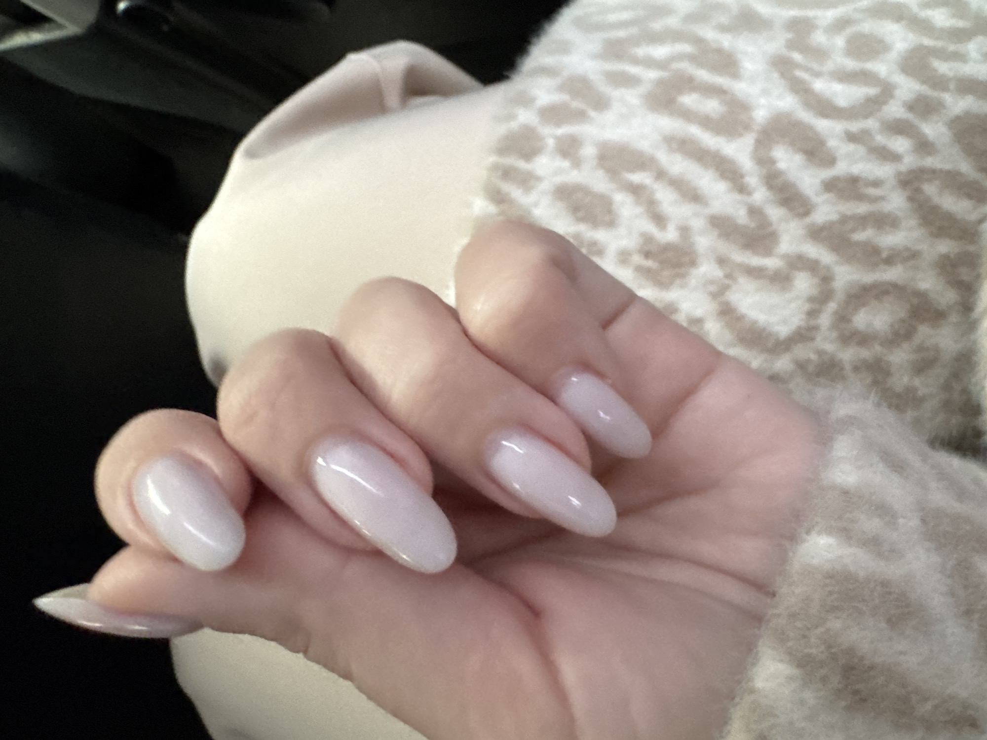 A hand is shown with long, almond-shaped nails painted a light pinkish-white color. The hand is positioned in front of a beige-colored surface with a blurry animal print pattern. Some black is visible in the upper-left corner. The overall lighting is soft. There is no indication this image was taken in Chaikovsky, Russia.