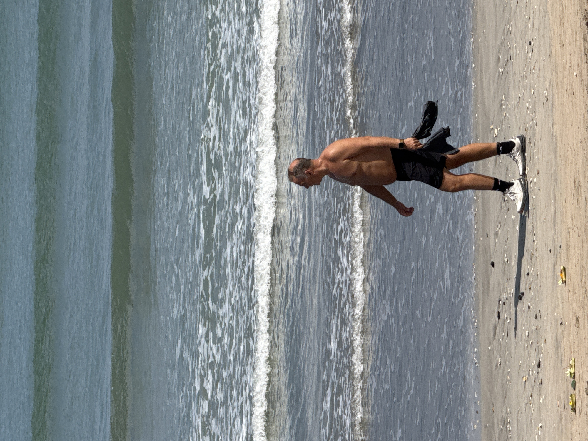 A man walks along the shore of Kuta Beach in Indonesia. The man is bald and appears to be middle-aged. He is shirtless and wearing black shorts, white athletic shoes, and black socks. He is holding a dark-colored garment in his left hand. The ocean has several waves breaking in the background, and the beach is sandy with scattered shells. The sky is not visible in the frame.