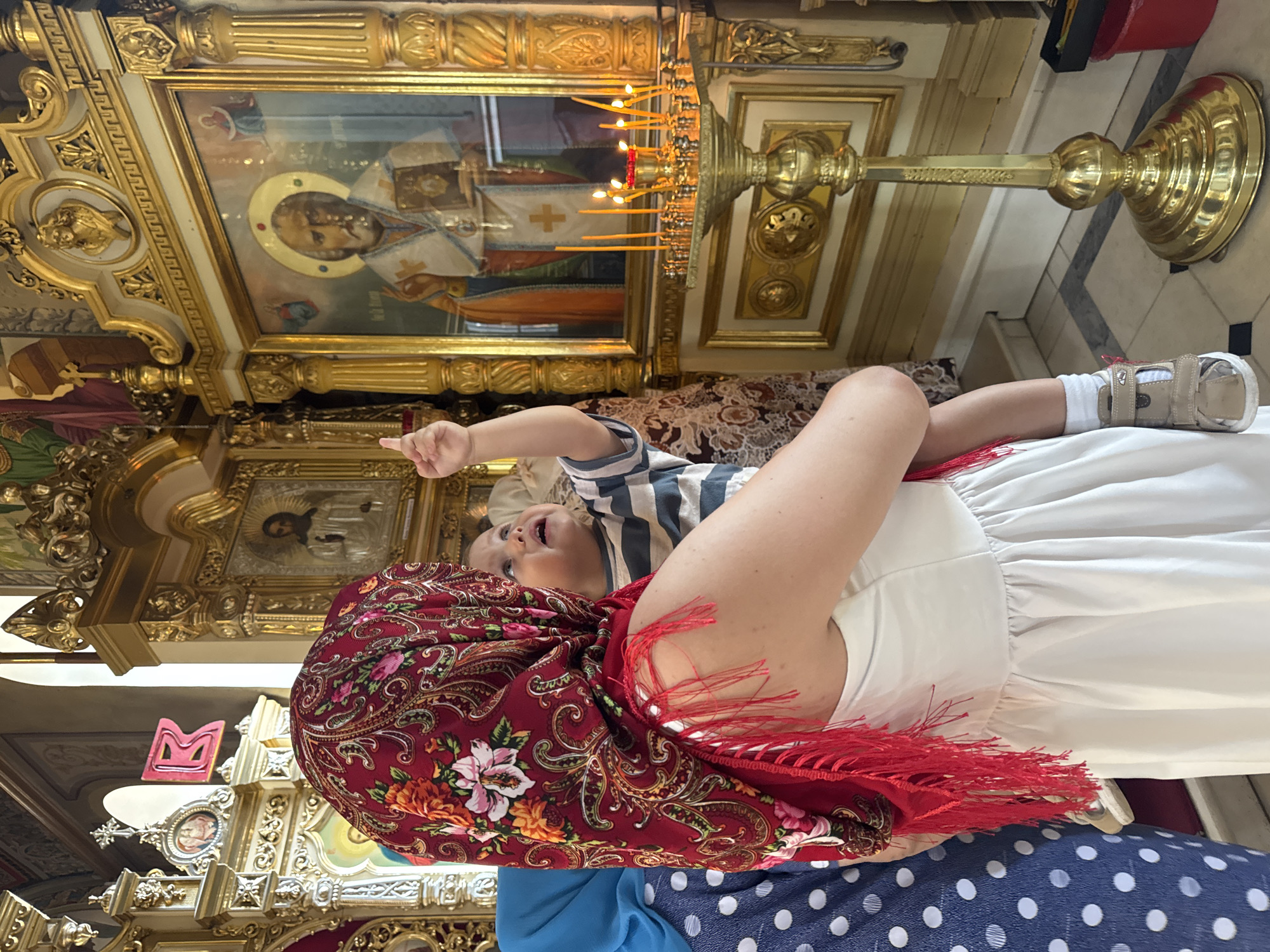 This image depicts a mother holding her young child in what appears to be an Orthodox church. The mother, wearing a red patterned headscarf and a white dress, is facing away from the camera, towards an iconostasis. The child, dressed in a striped shirt and a diaper, is being held by the mother and is looking up, pointing at something in the distance. A woman in a blue polka dot dress is partially visible to the left. The background features ornate religious artwork, golden fixtures, and lit candles. The scene conveys a sense of reverence and family. There is no information in the image that indicates the location is the city of Tolyatti, Russia.