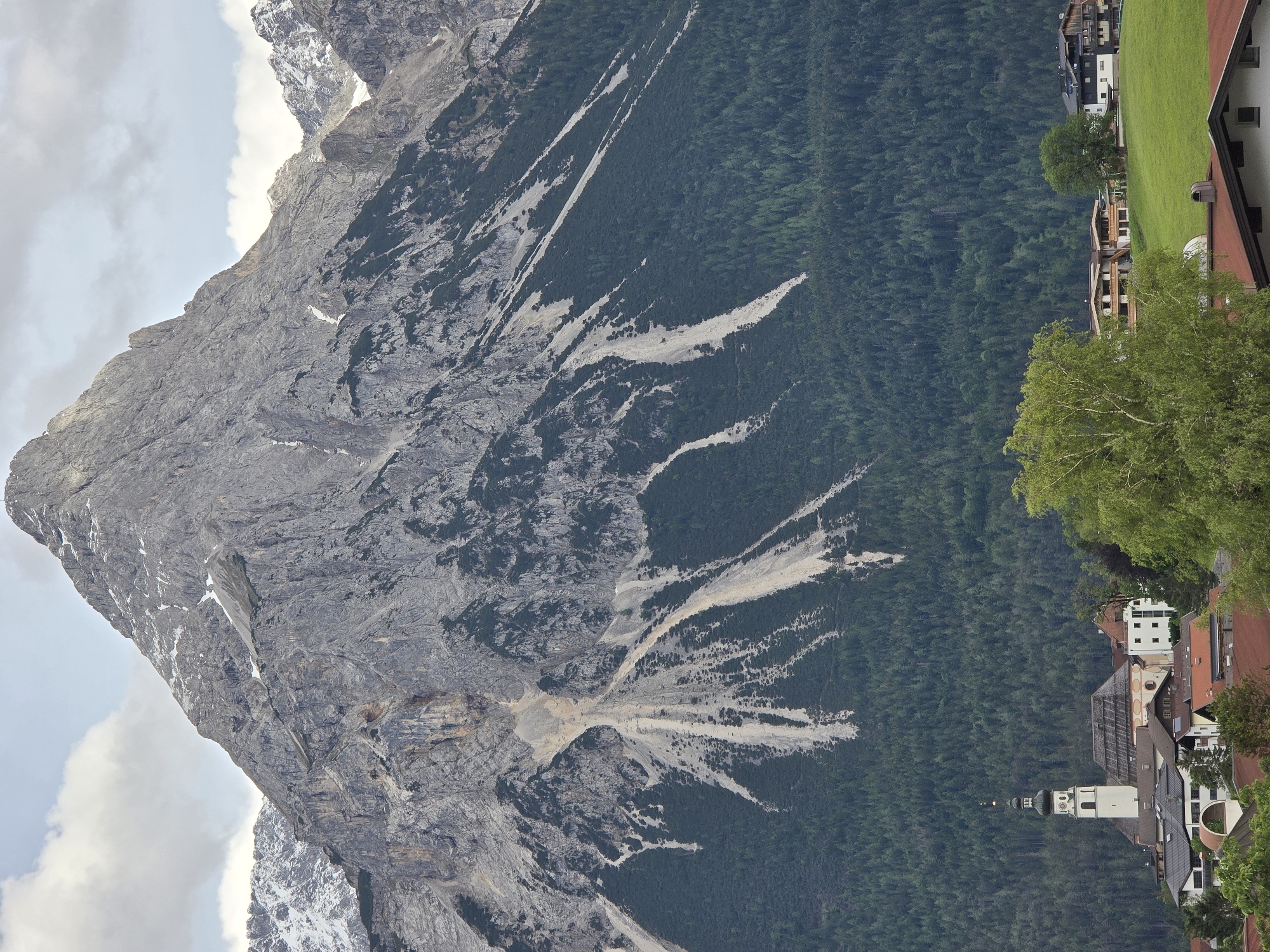 The image depicts a view of Lermoos, Austria, with a prominent mountain range in the background. In the foreground, a village is nestled at the base of the mountains. A white church with a distinctive black spire and clock face stands out among the buildings. The village consists of various houses with red-tiled roofs and some modern structures. The mountains are characterized by steep, rocky slopes with patches of snow near the summit, and dense green forests covering the lower sections. The sky is partly cloudy with a mix of white and gray clouds. The scene captures a serene, mountainous landscape with a traditional European village.