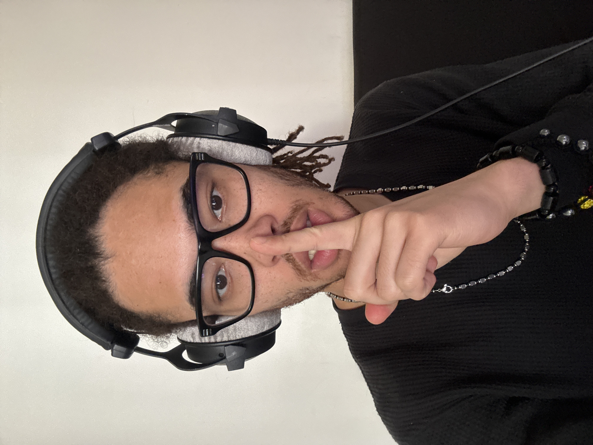 A person with brown skin, glasses, and headphones is holding a finger to their lips. They are wearing a black shirt and necklaces. The background is a plain white wall on the left and a black wall on the right. The location is not identifiable as Paris, France based on the image.