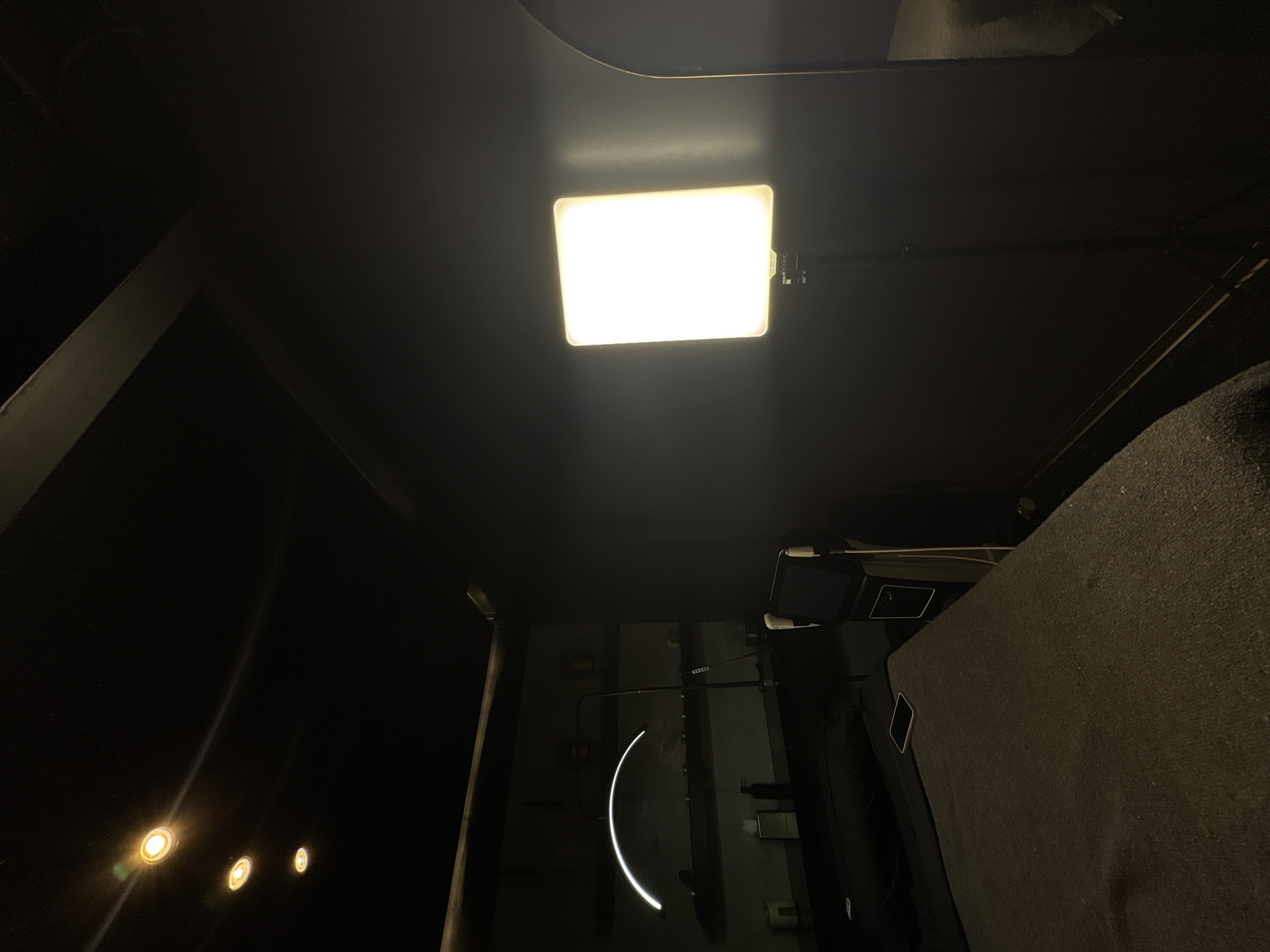 The image shows an interior space at night with artificial lighting. A rectangular LED panel light is mounted centrally on a dark ceiling. Further back, there is some lighting in the bottom left corner. On the right, a dark upholstered surface is visible, and toward the bottom a vertical wall runs upwards from the bottom edge to the middle. Further back a curved tube light illuminates the vertical wall. A monitor is fixed on the wall. I have no information to identify the specific location as Ciudad López Mateos, Mexico.