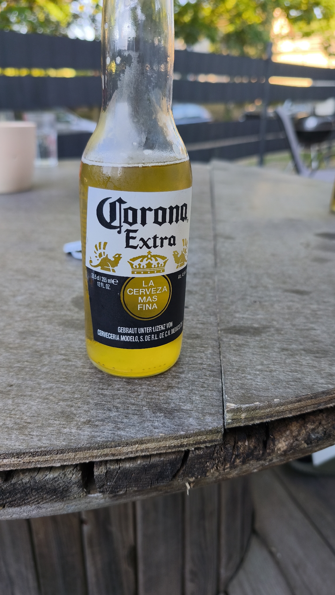 An outdoor, eye-level shot focuses on a clear glass bottle of Corona Extra beer, half-filled with a yellow liquid, resting on a weathered wooden table. The upper part of the bottle shows condensation, indicating it is cold. The label, predominantly black and yellow, clearly displays "Corona Extra" at the top in a distinctive font, with a golden crown and other emblems below. Other visible text includes "35.5 cl / 355 ml e", "12 FL. OZ.", "LA CERVEZA MAS FINA" (The Finest Beer), "GEBRAUT UNTER LIZENZ VON", and "CERVECERIA MODELO, S. DE R.L. DE C.V. MEXICO". Part of "ALC 4.5%" is also visible.
The wooden table appears rustic and worn, with visible grain and some damage or rough edges, suggesting an outdoor setting like a patio or deck. In the blurred background, green foliage from trees or bushes is visible, along with a dark fence or railing and indistinct shapes that could be outdoor furniture or structures. The natural lighting suggests it is daytime, possibly late afternoon given the soft, warm tones in the background. No people are visible, and no specific activity is taking place, though the beer bottle implies a moment of relaxation.