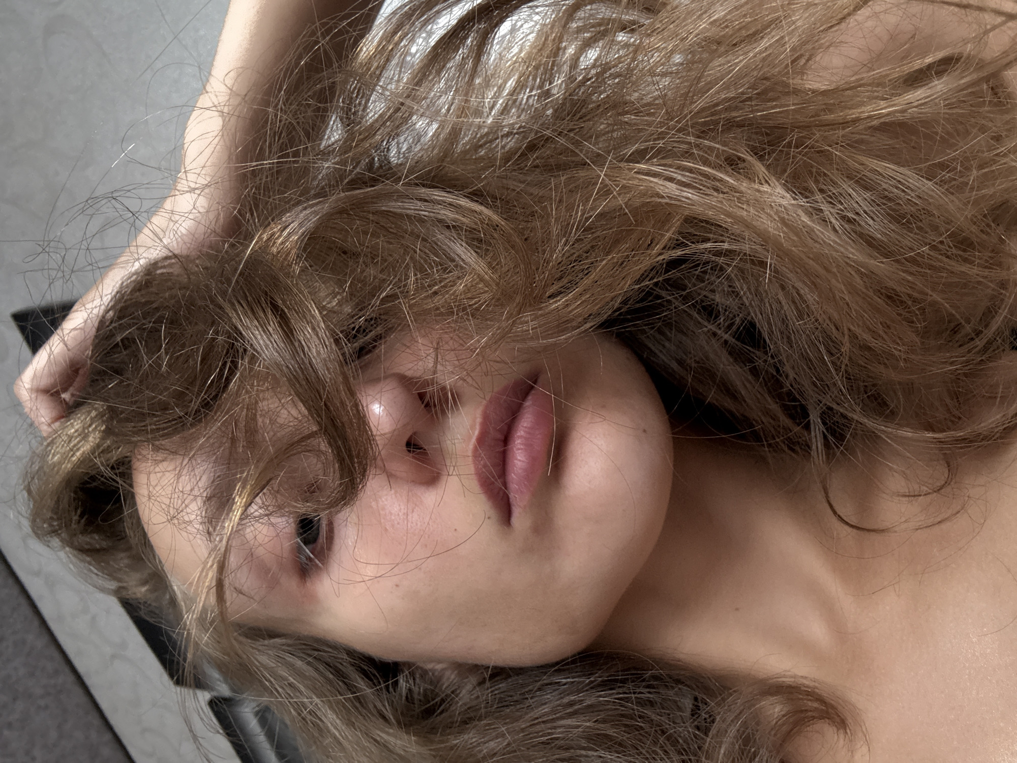 The image is a close-up, low-angle shot of a young woman's face and upper body, taken indoors. Her right hand is in her hair, pulling it back from her face. Her hair is long and a light brown color with blonde highlights, and it appears somewhat messy and windswept. Her face is partially obscured by her hair, but her lips are visible, slightly parted. Her skin is fair, with some freckles visible on her cheek. Her eyes are dark and partially visible. The background is blurred and appears to be a neutral-colored wall or surface. There is no indication of the location being in Kuyeda, Russia.