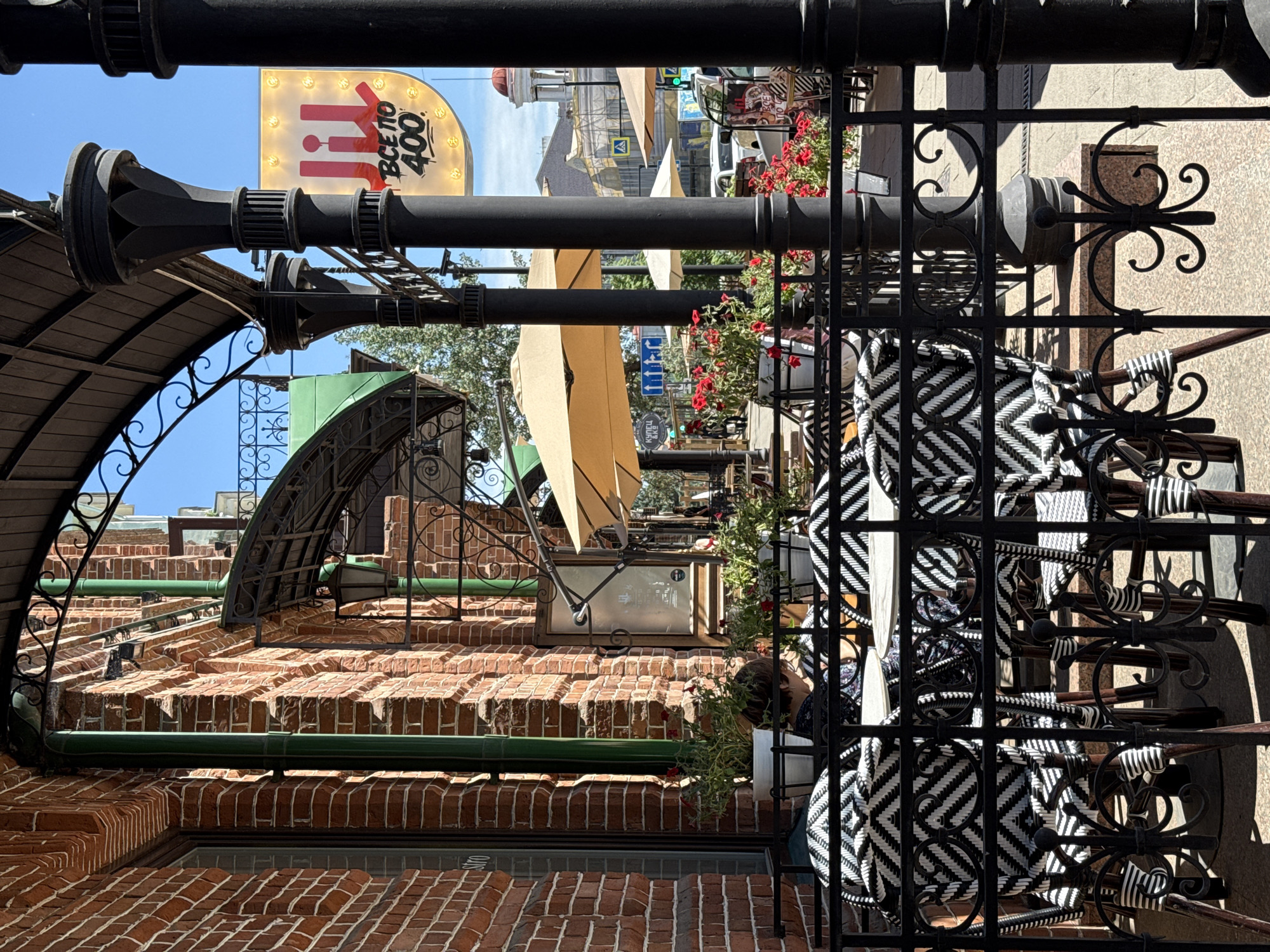 The image shows an outdoor seating area of a cafe or restaurant on a sunny day. In the foreground, there are black metal chairs with black and white patterned upholstery, arranged around tables. A black metal fence with decorative elements separates the seating area from the sidewalk. Behind the fence, there are potted plants with red flowers. A person is seated at one of the tables, facing away from the camera and looking down at something on the table.
The cafe appears to be located on a street with brick buildings. In the background, there are other outdoor seating areas with umbrellas, and pedestrian traffic. A bright yellow sign with red lettering and a stylized "JIJI" logo is visible in the background. The sign also has the text "ВСЁ ПО 400" which translates to "Everything for 400." This likely indicates a price point or special offer. Further in the background, architectural elements of buildings, including a domed structure, are visible. The overall setting suggests a European-style streetscape, potentially in a city center. Based on the prominent "JIJI" sign with Russian text, the location is identified as the JIJI store located at 35 Vzlyotnaya Street, Krasnoyarsk, Russia. The image was taken from the perspective of a pedestrian on the street, looking into the cafe's outdoor seating area.