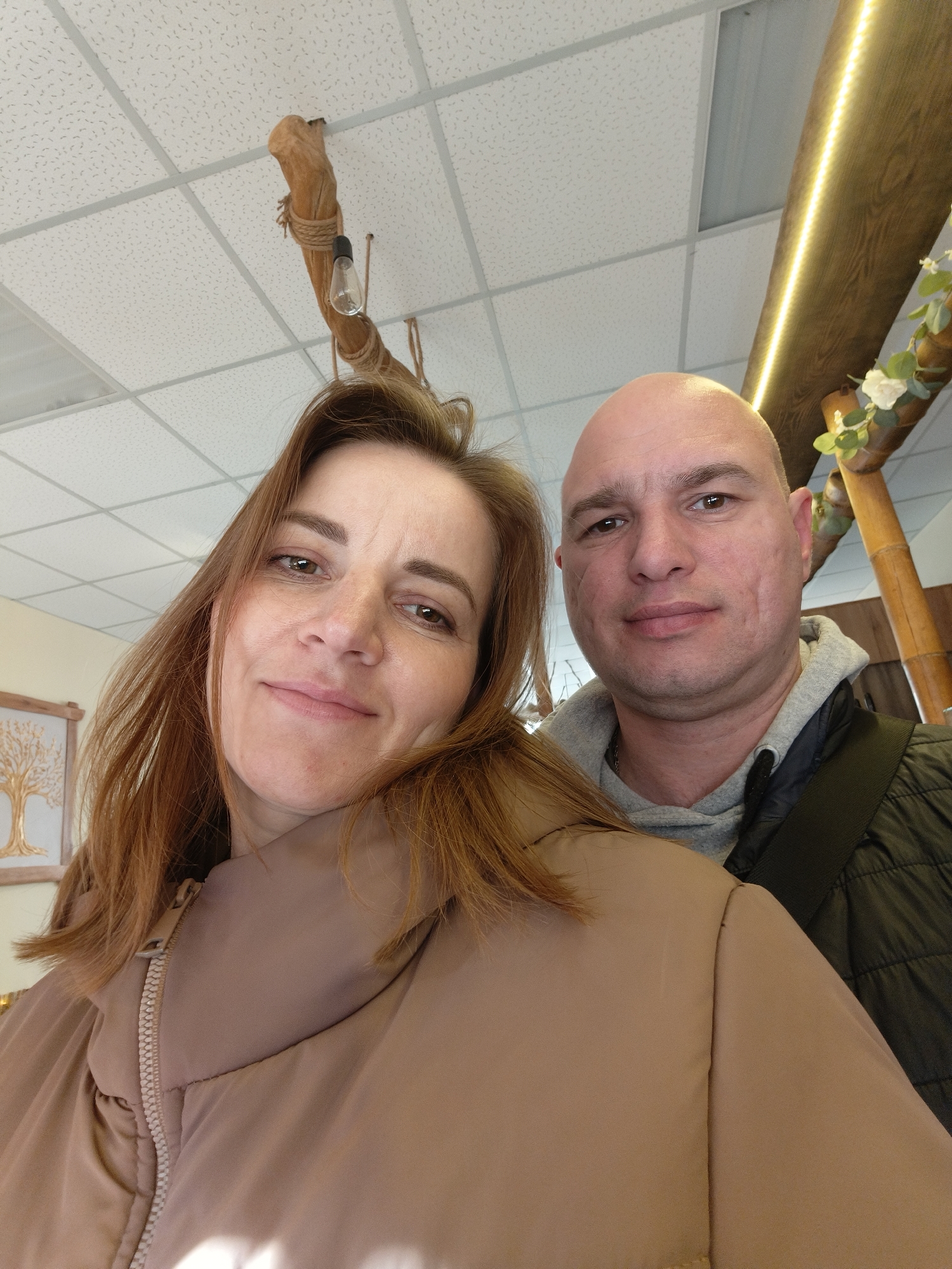 This is an eye-level indoor selfie featuring a woman and a man. The setting appears to be a commercial or public space, possibly a cafe or restaurant, given the decor and ceiling type. The location context is Kryvyi Rih, Ukraine.
The woman, positioned in the foreground and slightly to the left, has shoulder-length reddish-brown hair and light brown eyes. She is smiling faintly and looking directly at the camera. She is wearing a light brown or beige puffer jacket. Her expression conveys a sense of pleasantness.
Behind and to the right of the woman is a man with a bald head and brown eyes. He has a neutral expression, looking directly at the camera. He is wearing a grey hooded sweatshirt under a dark, possibly black, quilted jacket. A dark strap from a bag is visible over his right shoulder.
The background features a white acoustic tile ceiling with a grid pattern. Unique decorative lighting elements are visible: on the left, a rustic light fixture made from a thick wooden branch wrapped with rope, from which a bare light bulb hangs down. On the right, a wooden beam adorned with artificial white flowers and green leaves features a strip of warm LED lighting along its length. In the far left background, a framed piece of artwork depicting a golden tree is partially visible.
The image captures a moment of two people posing together, likely for a selfie, in an inviting indoor environment. There is no visible text in the image.