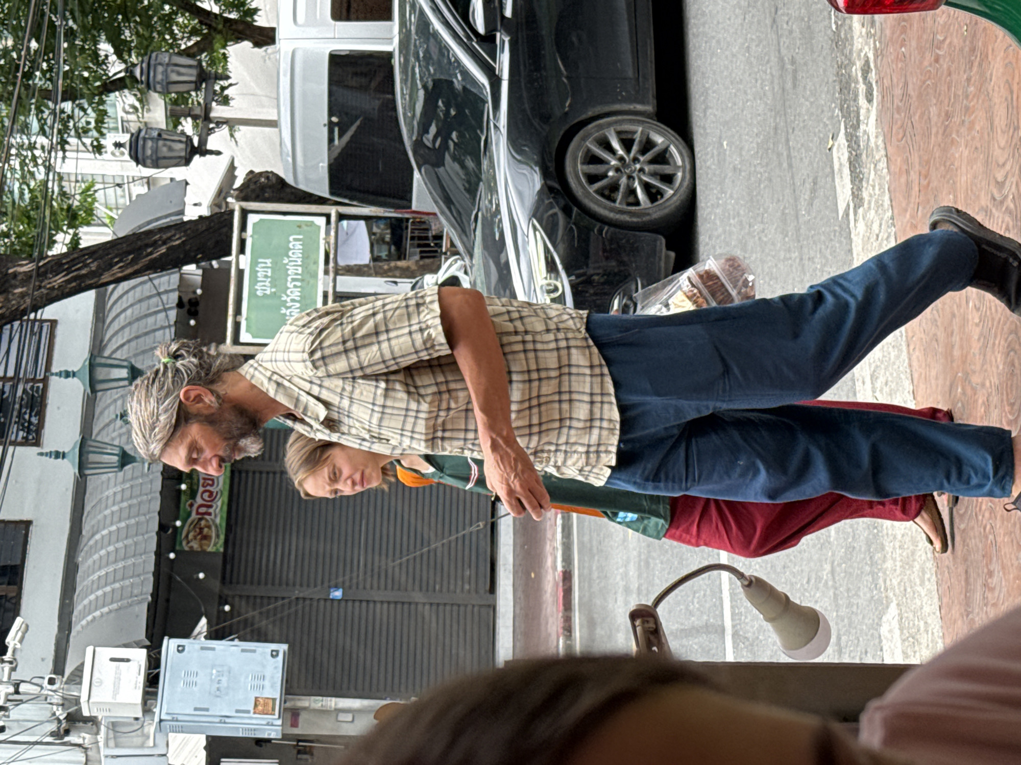The image depicts a street scene in Bangkok, Thailand. In the foreground, a man with graying hair pulled into a bun and a beard walks towards the viewer's right, wearing a plaid shirt and blue pants. He is holding a clear plastic bag containing food items. To his left, a woman with blonde hair walks in the same direction, wearing a green and orange shirt and red pants. She is looking to her left. Behind them, a black car is parked on the street, and a white van is visible further back. The environment includes a sidewalk with a patterned surface, streetlights with ornate fixtures, electrical boxes mounted on a pole, and a building with a curved awning. A green sign with Thai text is also visible. The overall impression is of a busy urban environment.