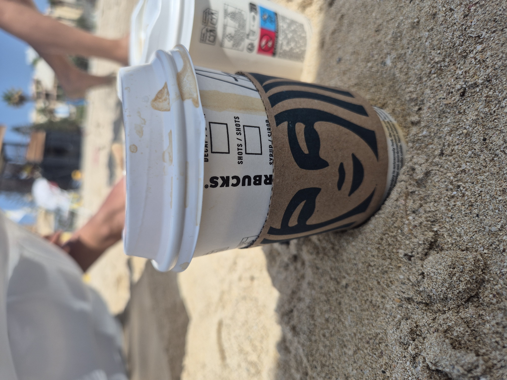 Two Starbucks coffee cups sit on a sandy beach. The cup in the foreground is white with a brown cardboard sleeve featuring the Starbucks logo. The lid has brown liquid stains. The cup behind it is also white. In the background, out of focus, are people walking on the beach, a palm tree, and buildings under a clear blue sky. The overall scene suggests a casual day at the beach in Birżebbuġa, Malta.