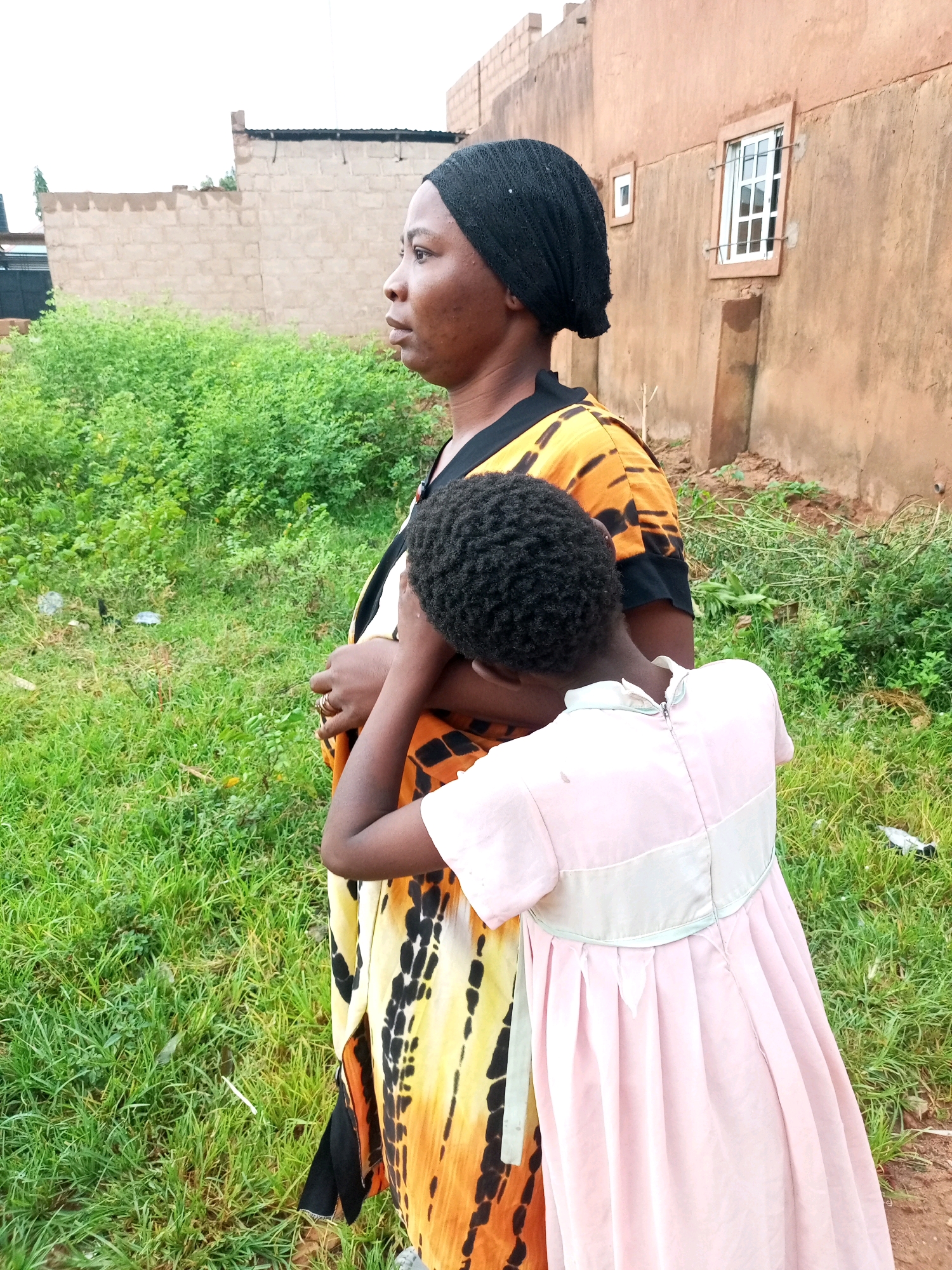 The image shows an outdoor scene in what appears to be a rural or semi-urban area, possibly in Tempure, Nigeria. In the foreground, a woman wearing a black headscarf and a brightly patterned orange and black dress is standing and looking to her left. She has her left arm around a young child who is facing away from the camera and is wearing a light pink dress. The child has its face turned towards the woman's chest, obscuring its facial features. The two subjects are standing on a patch of green grass and dirt. In the background, there is a building with a textured, earthy-colored wall and a white-framed window. To the left of the building, there is a section of a less finished wall made of concrete blocks, and beyond that, dense green foliage. The sky is overcast and a pale gray.