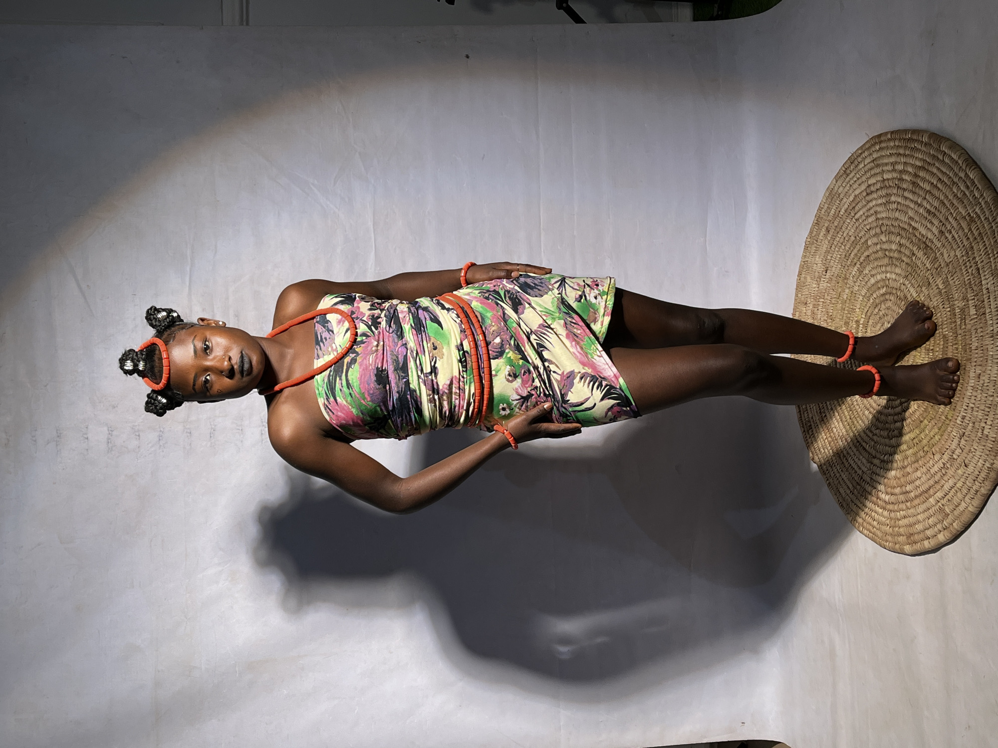 A young Black woman stands in front of a grey backdrop, her body angled slightly to the right, looking directly at the camera. She has dark skin and her hair is styled in two prominent buns on either side of her head, decorated with orange beads. She wears a strapless dress with a vibrant floral pattern in shades of green, pink, and yellow, cinched at the waist. Orange beaded jewelry adorns her neck, wrists, and ankles. Her makeup includes dark lipstick and eyeliner. She stands barefoot on a round, woven mat. A large, soft shadow of a person is cast on the backdrop to her left. The background appears to be a studio setting, with soft lighting creating a gentle gradient on the backdrop and a spotlight effect in the upper right quadrant. The scene has no discernible elements that would place it in Barakin Akawo, Nigeria.