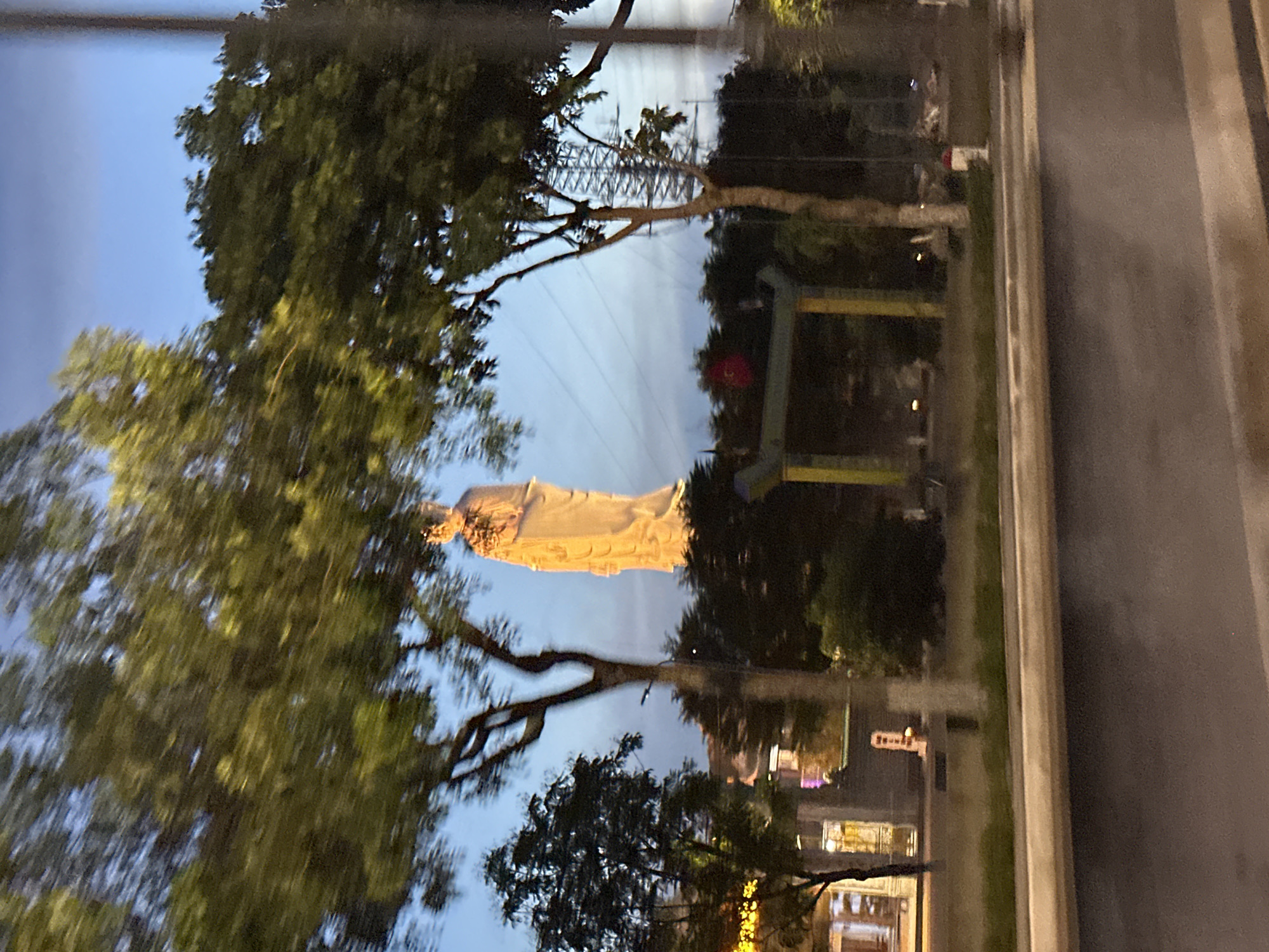 The image captures a serene evening scene in Đà Nẵng, Vietnam, featuring a towering white statue of Guanyin, the Buddhist goddess of mercy, illuminated against a twilight sky. The statue stands tall and majestic, adorned in traditional robes, with her hands held in a gesture of blessing. Surrounding the statue are lush green trees, their branches reaching towards the sky, adding a touch of natural beauty to the urban landscape. In the foreground, a road with a slight incline leads the eye towards the statue, with the blurred motion of vehicles suggesting the bustling city life that continues around this peaceful sanctuary. A traditional yellow gate structure is visible in the middle ground, hinting at the architectural style of the region. The overall ambiance is one of tranquility and reverence, a testament to the spiritual significance of the statue and its place within the city.